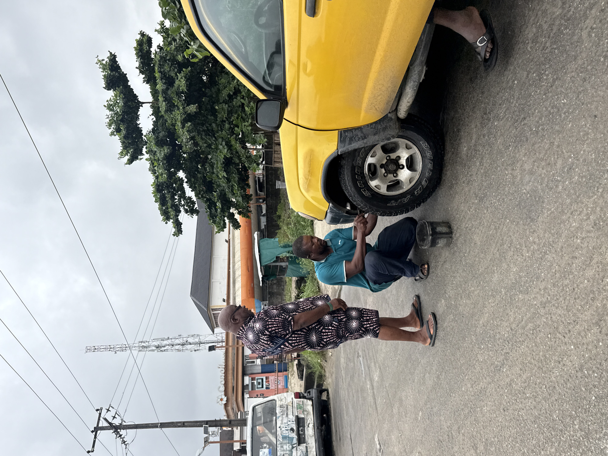In Ado, Nigeria, on an overcast day, two men are attending to a yellow SUV parked on a paved street. The man on the left, wearing a patterned shirt and shorts with black flip-flops, stands observing. The man on the right, dressed in a teal polo shirt and dark trousers with black flip-flops, is crouched down, appearing to work on the front tire of the SUV with a tool and a small metal bucket nearby. In the background, a white van is parked, and a business with orange and white signage, possibly a gas station, is visible. Utility poles and a distant cell tower are also present. Trees with dense green foliage are on the right side of the image.