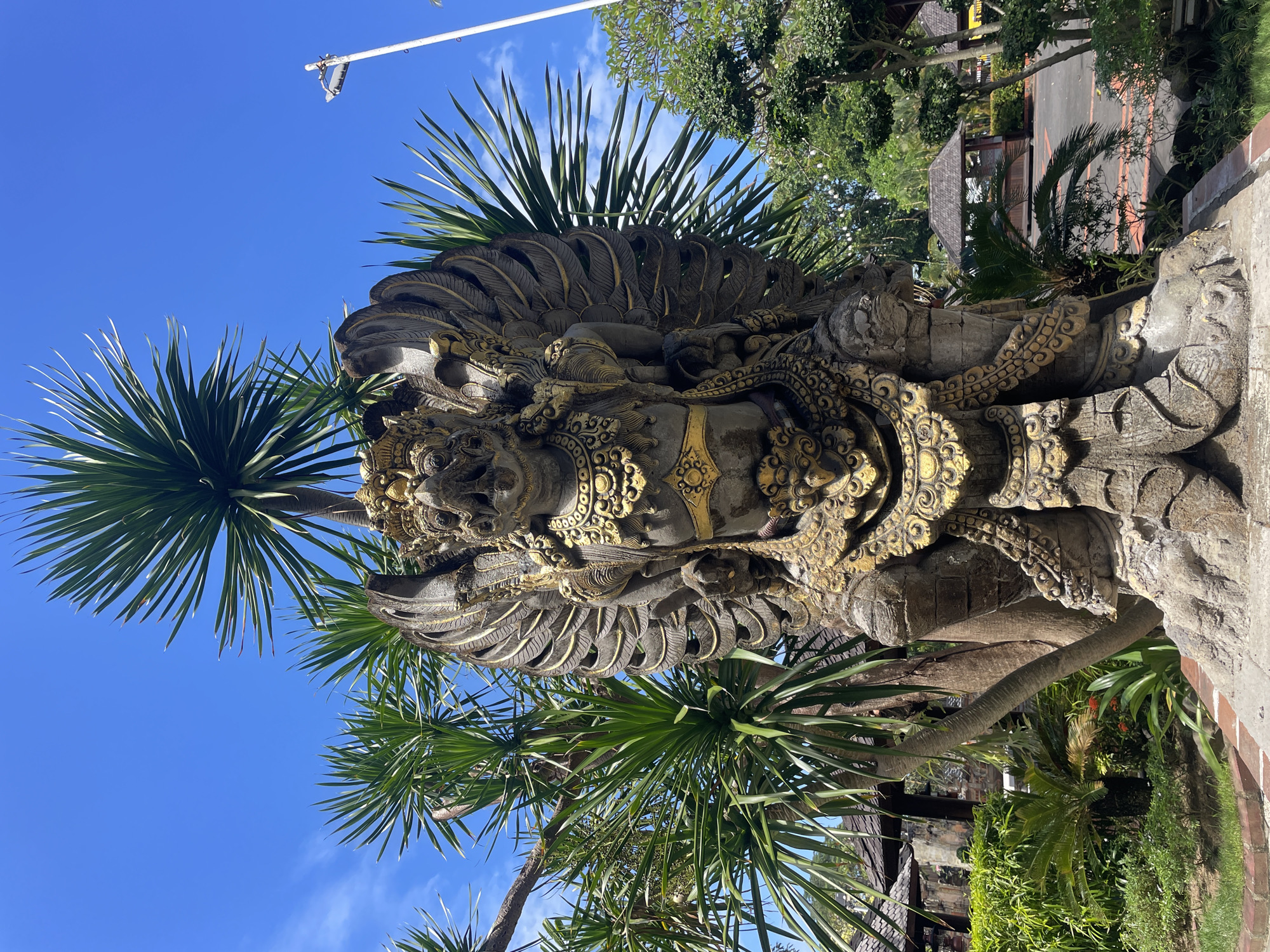 The image shows a highly detailed, ornate stone sculpture in Denpasar, Indonesia. The sculpture is primarily gray with golden accents, depicting a mythological figure adorned with intricate carvings. Palm trees surround the sculpture, and a clear blue sky is visible in the background. In the upper-left corner, a utility pole with attached wires is present. The setting appears to be a garden or park area, with additional greenery visible. In the background, structures suggest buildings or pavilions characteristic of the location.