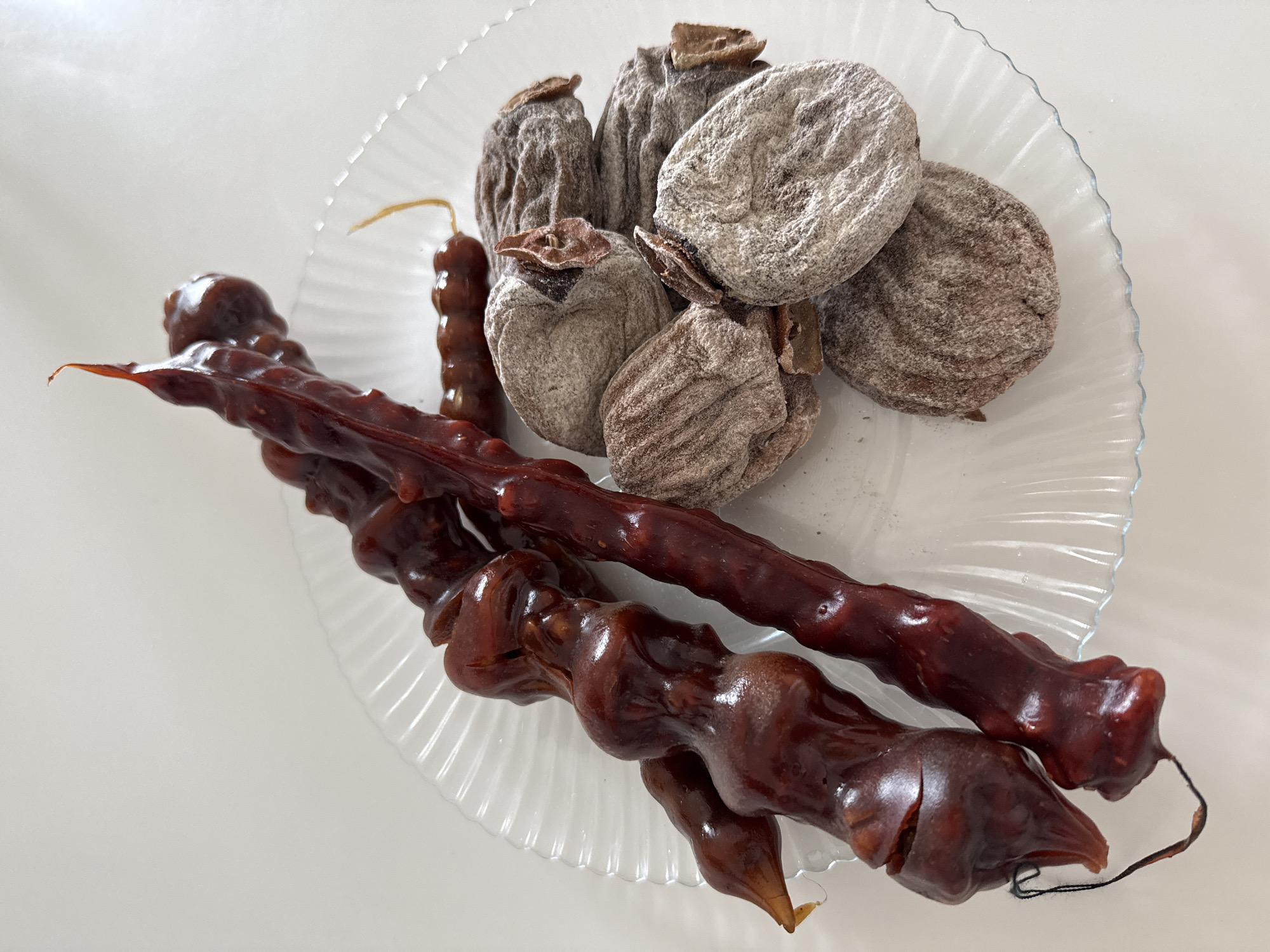 A glass plate with scalloped edges sits on a white surface. On the plate are two long, dark brown Churchkhela, arranged parallel to each other, and a pile of five round, grayish-brown dried persimmons.