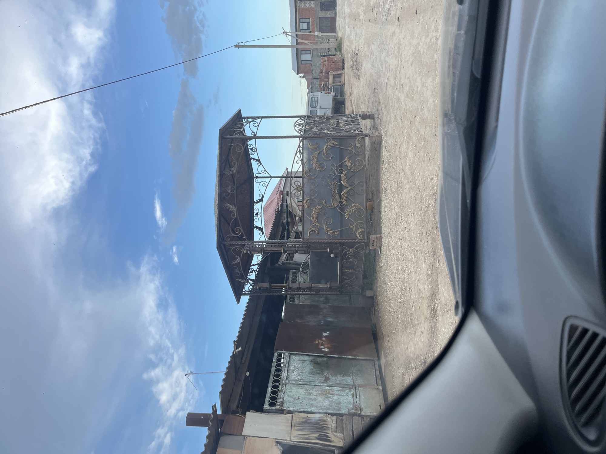 The image shows a view from inside a vehicle, likely a car, looking out onto a street in Kaspiysk, Russia. The scene includes a metal gazebo with ornate wrought iron detailing, situated near a weathered building with metal doors and corrugated roofing. The surrounding environment consists of a dirt road, power lines, and a blue sky with scattered clouds. In the background, there are glimpses of other buildings, including one constructed from brick and a white vehicle. The vehicle from which the photo is taken features a gray interior.