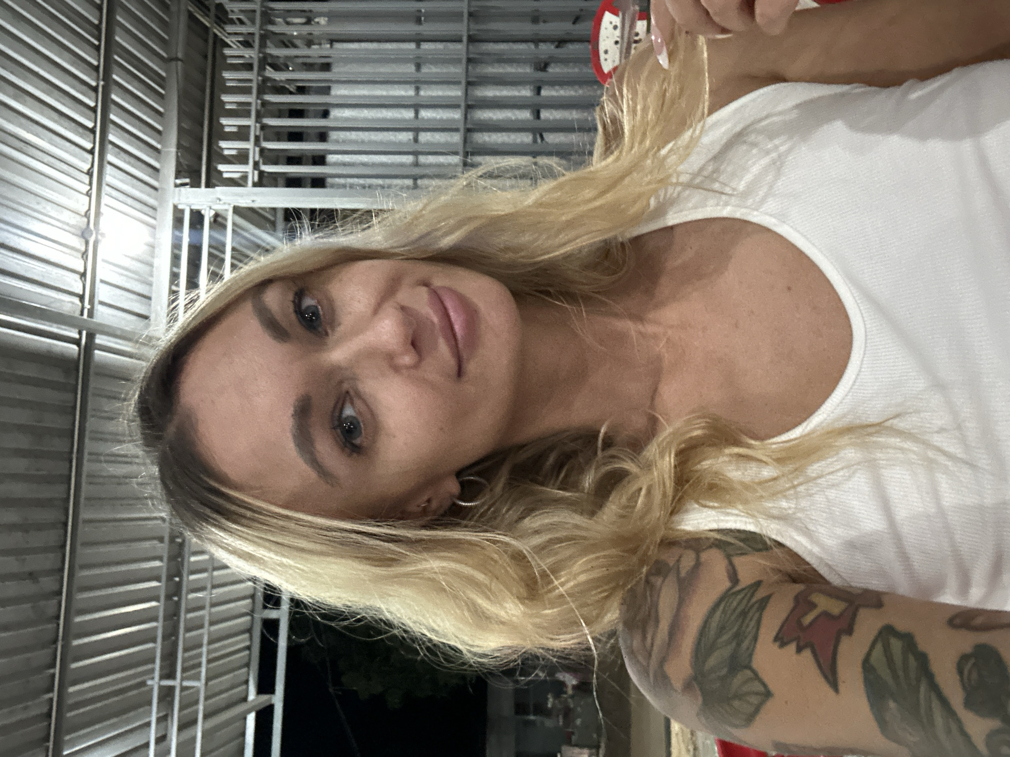 A blonde female is centered in a selfie-style portrait, taken at night in Phu Thuy Ward, Vietnam. She has long, wavy blonde hair parted to her left, blue eyes, and is smiling faintly with slightly parted lips. She wears a white ribbed tank top. Her left arm is heavily tattooed with a partial sleeve, featuring leafy designs and a distinct red and yellow emblem, possibly a crest or stylized letter, on the bicep. She has small silver hoop earrings on both ears and an upper helix piercing on her left ear. Her right hand, with white French-tipped nails, is visible at the lower right, appearing to lightly touch her hair.
The background depicts an open-air or semi-enclosed structure. Above her, a corrugated metal roof with visible support beams is brightly illuminated. To her right, a vertical corrugated metal wall or partition is present. The area beyond the immediate structure is dark, with a faint silhouette of trees visible on the far left. A glimpse of red and white objects, possibly chairs or furniture, is seen in the lower right background. The scene is well-lit, with direct illumination on the subject and the overhead structure.