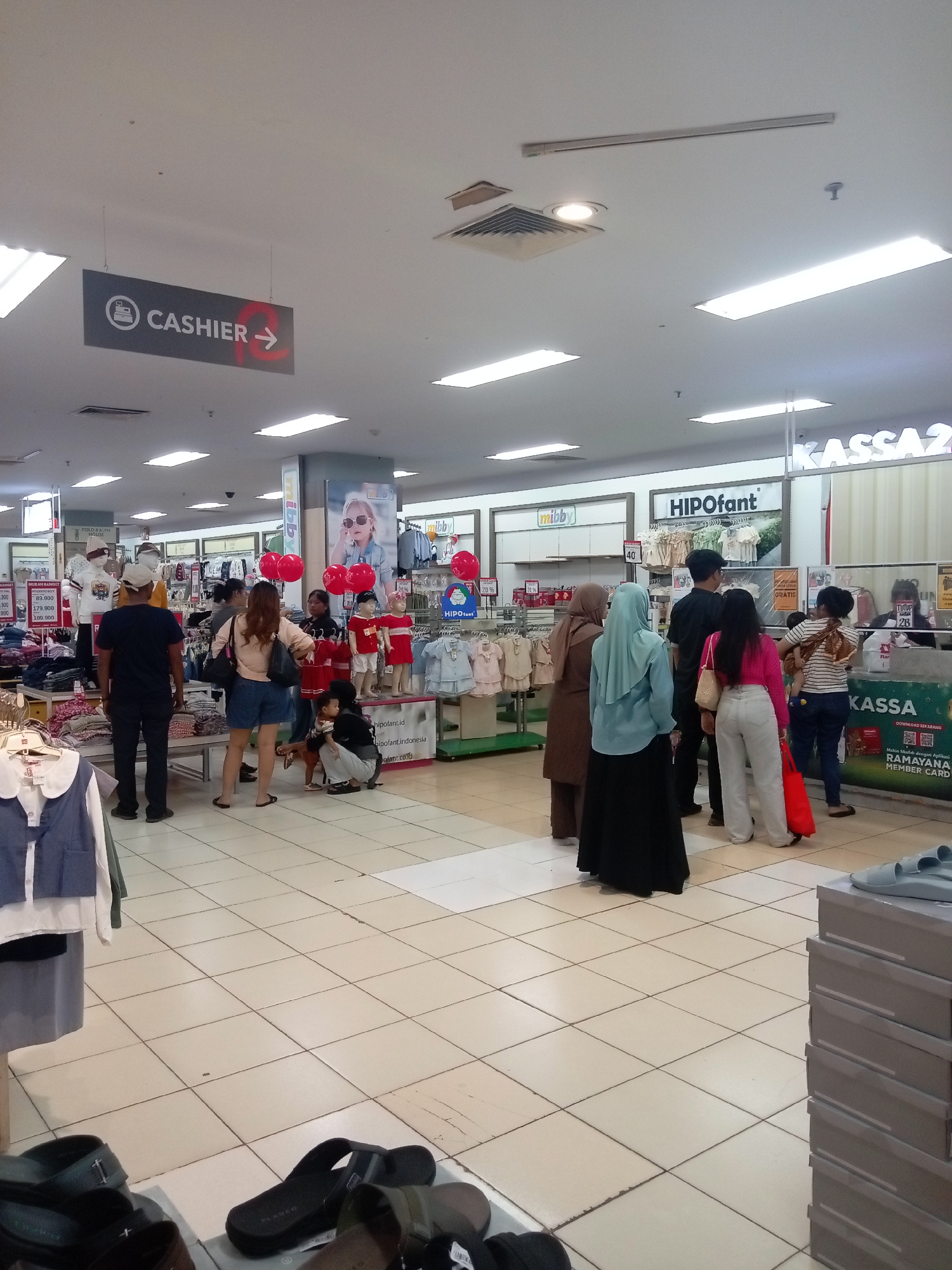 The image captures a busy scene inside a department store, likely in Batam, Indonesia, given the context of the "Ramayana" branding visible. In the foreground, a stack of shoe boxes with light blue sandals on top is visible on the right, and various pairs of sandals are laid out on the floor on the left. The main area of the store is filled with people browsing and queuing. A prominent "CASHIER" sign with an arrow pointing to the right is suspended from the ceiling.
Several groups of people are dispersed throughout the store. Near the cashier area on the right, a group of individuals, including at least one cashier, are attending to customers. Further back, another group of shoppers is gathered around displays of children's clothing, some of which are adorned with red balloons. Mannequins dressed in children's wear are also present. The store environment features tiled floors, fluorescent lighting panels on the ceiling, and promotional banners and signage for brands like "HIPOfant" and "mibby." The overall impression is that of a commercial retail space with moderate customer traffic.