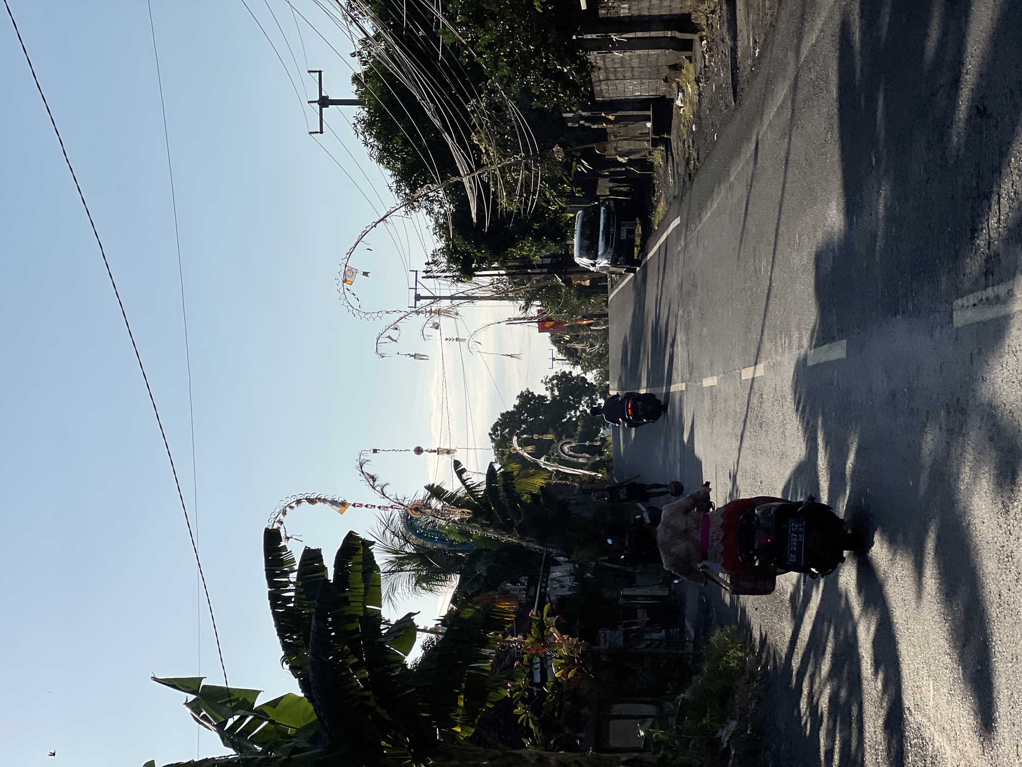The image depicts a street scene in the city of Kubu, Indonesia, bathed in the warm light of what appears to be early morning or late afternoon. The road, asphalted and marked with a white line down the center and a dashed white line on the right edge, slopes gently upwards. On the left side of the road, there are buildings with visible roofs, and lush green banana trees with broad leaves casting long shadows across the pavement. Traditional Balinese decorations, known as "penjor," adorn poles along the roadside, featuring curved bamboo structures decorated with fabric and other ornaments, suggesting a cultural or festive event.
Several people are present, primarily riding motorcycles. In the foreground, a person is riding a black scooter, viewed from behind. This individual is wearing a patterned top and a bright pink sarong or skirt. Further up the road, another person is riding a dark-colored motorcycle. The scene is also populated by utility poles with a dense network of electrical wires, typical of a developed area. On the right side of the road, a light-colored SUV is parked, and a stone wall with vertical posts runs along the property line, separating the road from what appears to be a residential or commercial area. The sky is clear and a pale blue.