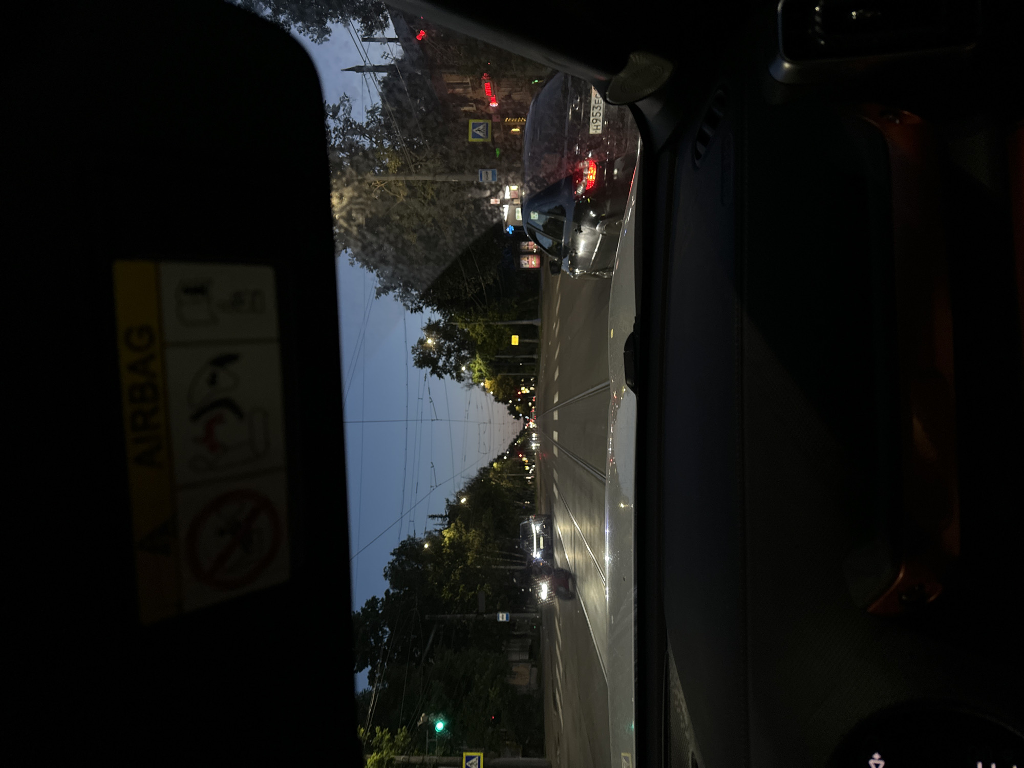 The image captures a view from inside a vehicle, likely a car, looking out through the windshield and side window onto a street scene in Nizhny Novgorod, Russia. The time of day appears to be dusk or early evening, as indicated by the dim lighting and the illuminated streetlights and vehicle lights. The weather seems to be clear, with a light blue sky visible in the distance.
The main subjects are the street, with its tram tracks running down the center, and the surrounding environment. Trees line the street, their foliage illuminated by streetlights. Several cars are visible, some stationary in what appears to be traffic and others moving. One car in the foreground has its license plate visible, reading "H953EC". Traffic lights are also present, with one green light visible. Pedestrian crossing signs and other traffic signs are also present.
The scene suggests a typical urban commute or a drive through the city during a low-light period. There are no people clearly visible within the frame, but their presence is implied by the occupied vehicles.
Notable details include the "AIRBAG" warning label on the interior of the vehicle, a common safety feature indicator. The tram tracks are a strong visual cue for the location, indicating a city with a tram system. The overall mood is calm and ordinary, depicting a moment in urban daily life. There is no explicit text that conveys a specific event or emotion, other than the standard vehicle warnings.