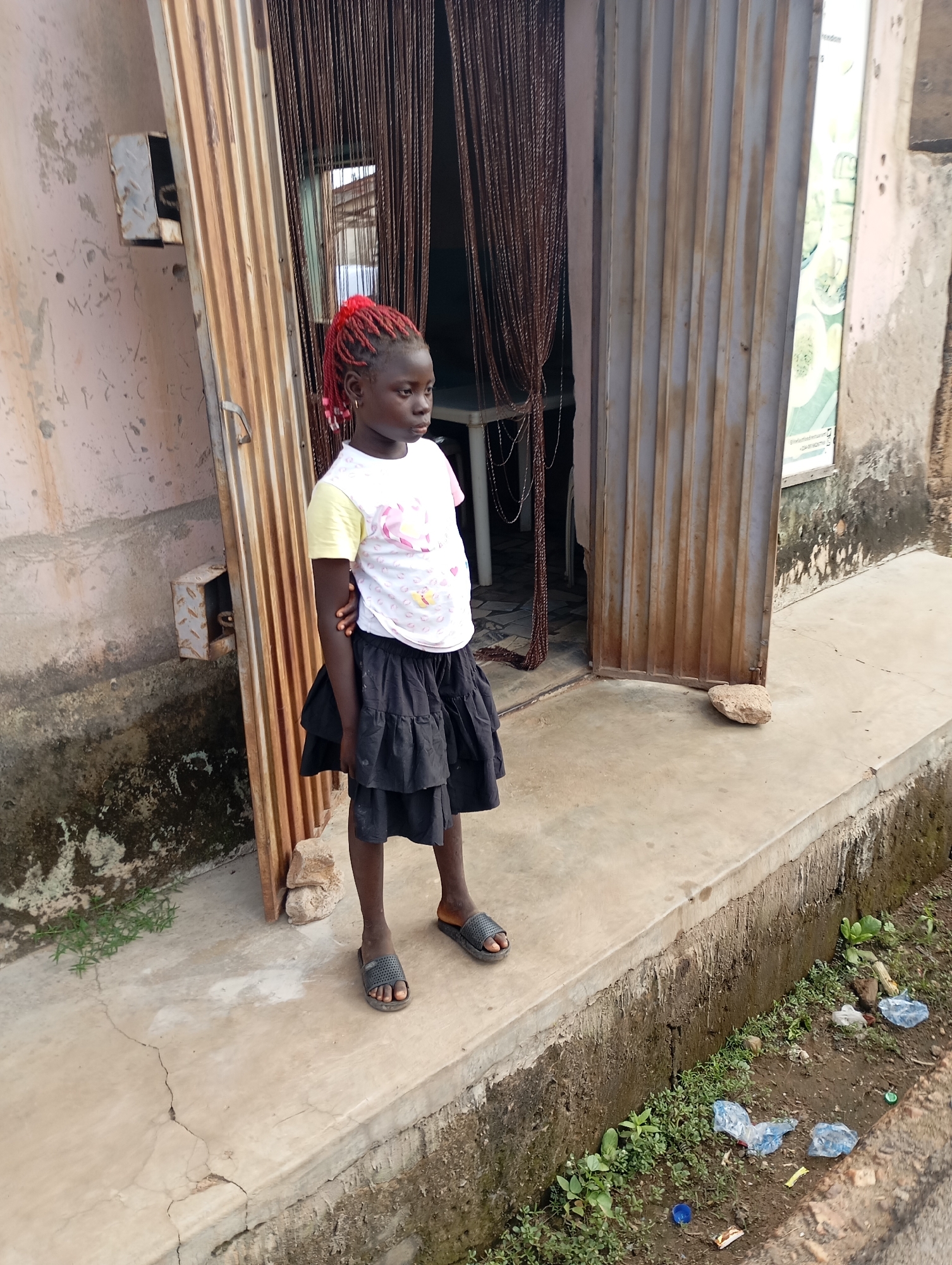 A young girl with bright red braided hair stands on a concrete sidewalk in front of a corrugated metal storefront. She wears a white t-shirt with a colorful graphic and a black ruffled skirt. Her feet are clad in dark gray slide sandals. The storefront is partially open, revealing a curtain of dark brown cords hanging inside, with a table visible behind them. To the left of the storefront, a rough, light-colored wall with a metal box attached is visible. To the right, the concrete sidewalk ends, dropping down to a dirty ditch filled with green plants and scattered plastic bottles and trash. The overall scene suggests an outdoor, possibly commercial, area within a city.