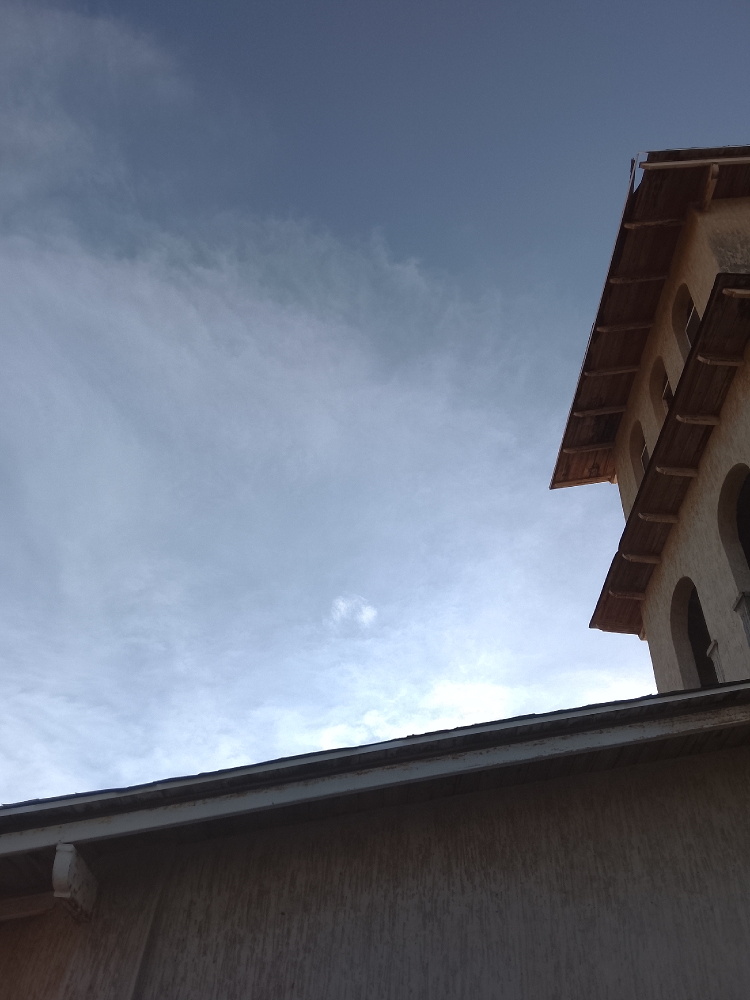 The image captures an upward-angled view of architectural details against a sky, set in Juan Griego, Venezuela. The majority of the frame is dominated by a soft blue-grey sky, indicating an overcast or partly cloudy day. Wispy, diffused cloud formations are present throughout the sky, with lighter areas suggesting indirect sunlight.
In the upper right portion of the image, the top section of a building is visible, angled upwards from the lower right corner. This building features a light-colored, textured facade, possibly stucco. It displays a series of arched openings or windows. A prominent architectural detail is its roofline, characterized by extended eaves supported by exposed wooden beams or corbels. This design suggests a traditional or Mediterranean influence. The roof material appears dark, possibly tiles or shingles, partially obscured by the overhanging eaves.
Running across the bottom of the frame and extending into the lower left is the roofline and upper wall of another structure. This building has a visible gutter system along its roof edge. Its wall also appears light-colored and textured, though less detailed than the building on the right.
The perspective is from a low vantage point, looking up towards the sky and the upper parts of these structures. No people or specific activities are visible in the scene. The lighting suggests it is either late morning or late afternoon, with the sun's direct rays being diffused by the clouds. There is no visible text in the image. The overall impression is one of calm and architectural observation under a muted sky.