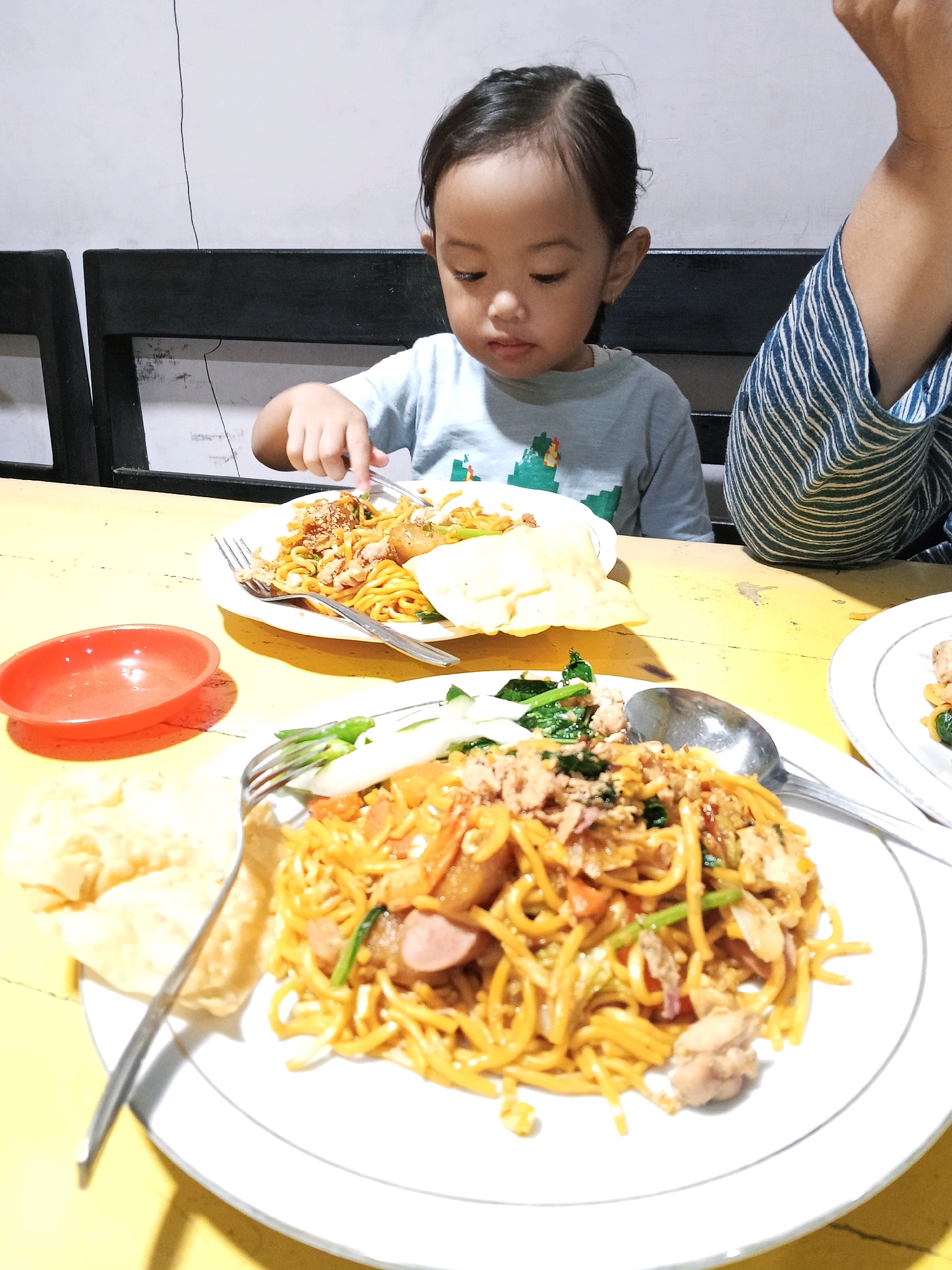 A young Indonesian child with dark hair is seated at a yellow wooden table, eating noodles. The child is wearing a light blue t-shirt with a graphic design and is holding a fork, engaged in eating. In front of the child is a white plate with noodles, crackers, and some green vegetables. To the child's left is a small red bowl. A second plate of noodles is in the foreground. The child is positioned between two dark wooden chairs, and the background appears to be a light-colored wall. The overall scene suggests a casual mealtime, possibly at a home or a local eatery in Blitar, Indonesia.