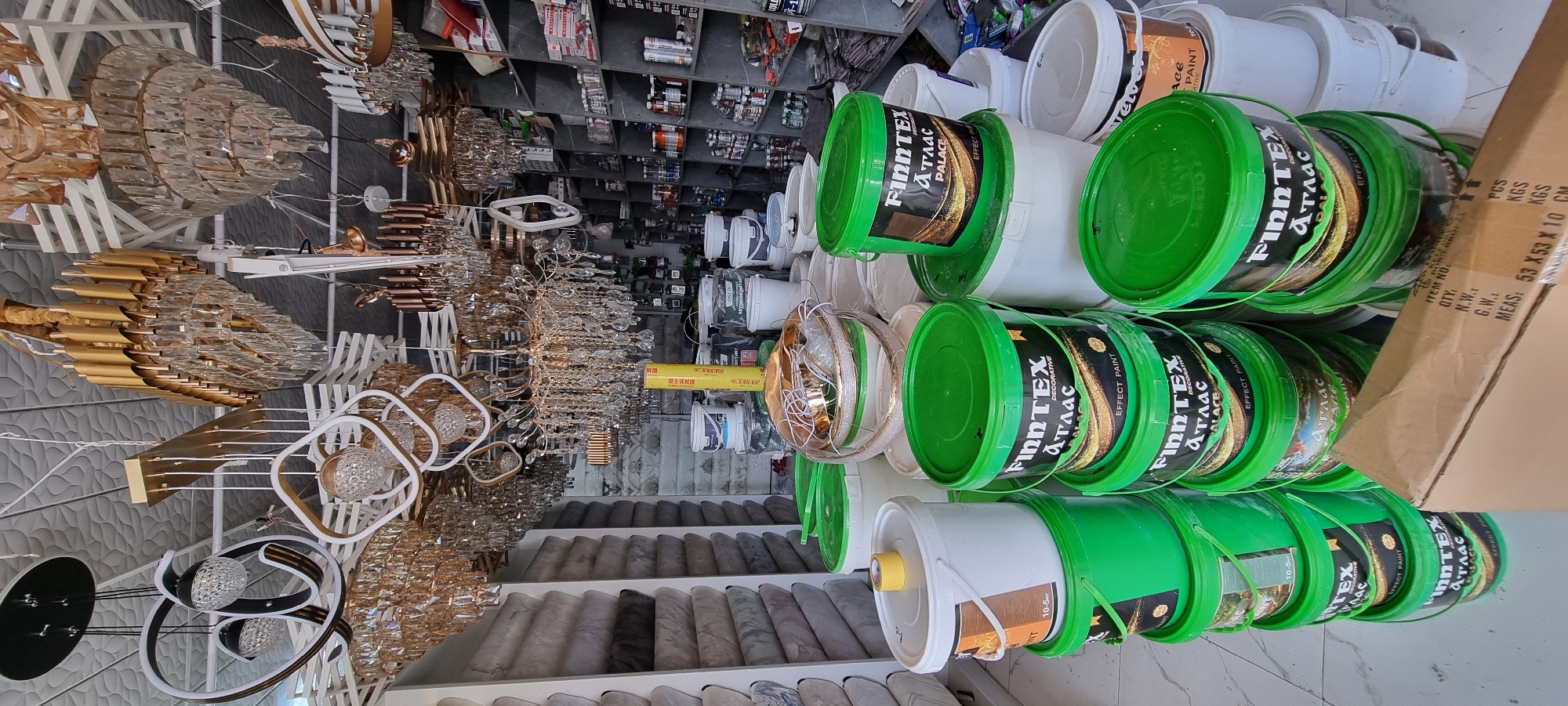 The image shows an interior view of a retail store selling home decor and building materials in Bokhtar, Tajikistan. In the foreground, several green and white buckets of paint are stacked, with prominent "FinnTex" branding. The buckets have labels indicating "Effect Paint" and "Palace". Behind the paint, various modern and ornate chandeliers are suspended from the ceiling. To the left, shelves are lined with rolls of what appears to be wallpaper. To the right, shelves are filled with numerous jars and bottles, likely containing paint or finishing products, as well as boxes of various goods. The overall impression is of a well-stocked store offering a range of interior design and renovation supplies.