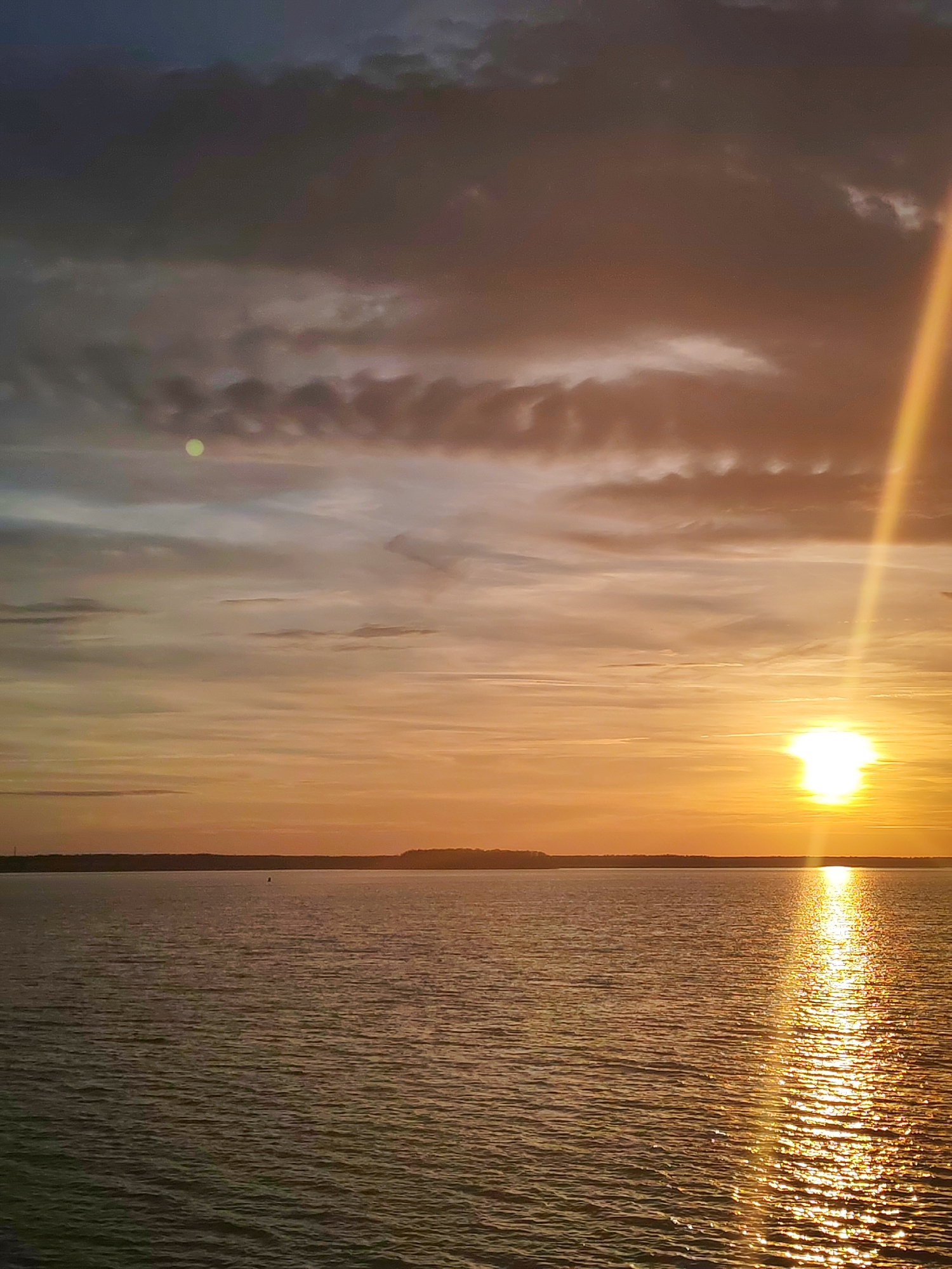 The image depicts a sunset over a body of water. The sky is filled with clouds, with varying shades of orange and grey. A bright sun is near the horizon, creating a reflection on the water's surface. In the distance, a strip of land is visible. There are no people visible in the image. The location is in the city of Unknown, Unknown.