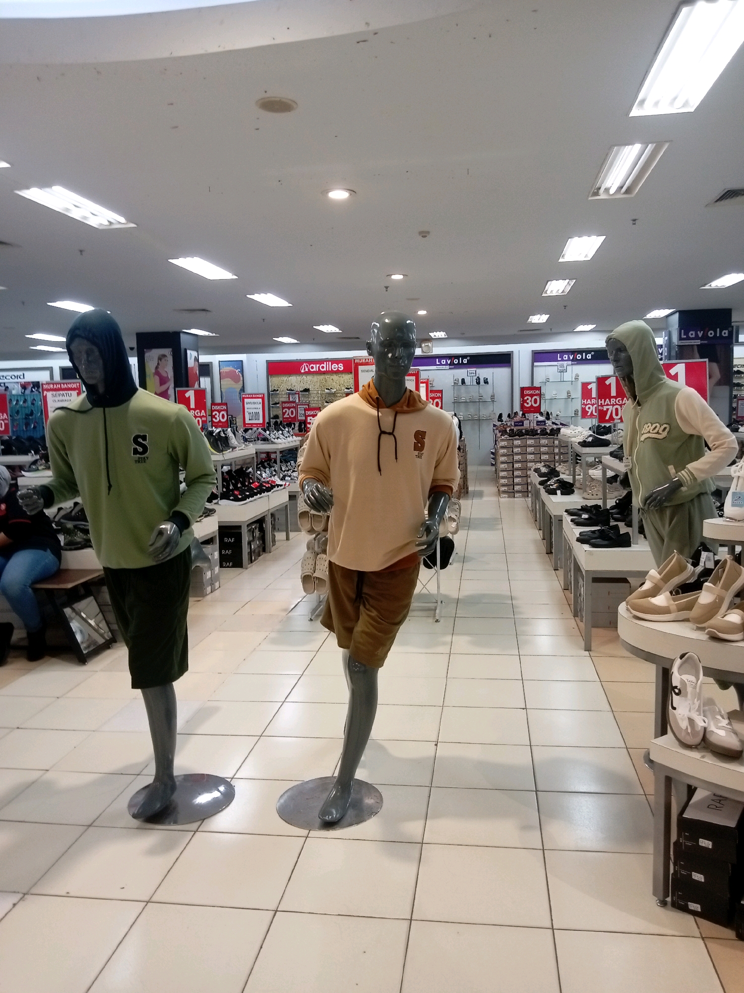 The image depicts an interior view of a shoe store, likely located in Batam, Indonesia, given the presence of Indonesian signage. In the foreground, two male mannequins are displayed, both positioned in running stances. The mannequin on the left wears a green hooded sweatshirt with a black hood and dark green shorts, featuring an "S" logo on the chest. The mannequin on the right is dressed in a light brown hooded sweatshirt and light brown shorts.
The store environment is brightly lit by fluorescent ceiling lights. Various shoe displays are visible in the background, arranged on white shelving units. Prominent signage for brands such as "Mardiles" and "Lavola" is present. Some signs indicate discounts, with text like "MURAH BANGET" (very cheap) and "DISKON" (discount). Stacks of shoe boxes are visible beneath some of the displays. A woman wearing blue jeans and a dark top is seated on a bench to the left of the frame, observing the merchandise. The floor is tiled with white square tiles.