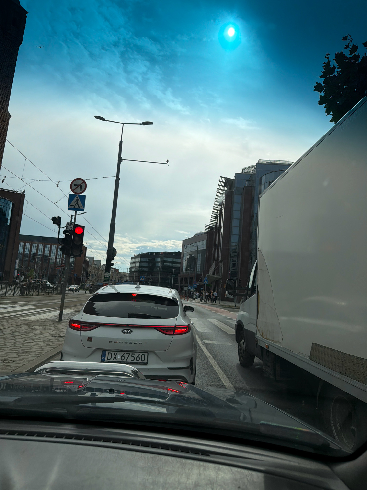 The image depicts a street scene in Wrocław, Poland, on a cloudy day with a bright, sun-like glare in the sky. A white Kia Proceed is stopped at a red traffic light, with its license plate showing "PL DX 67560". A large white truck is next to it, obscuring part of the view. In the background, modern buildings with glass facades line the street. Traffic lights, street lamps, and pedestrian crossing signals are visible. A no-turn sign is also present. People are seen walking on the sidewalk. The overall impression is an urban traffic intersection.
