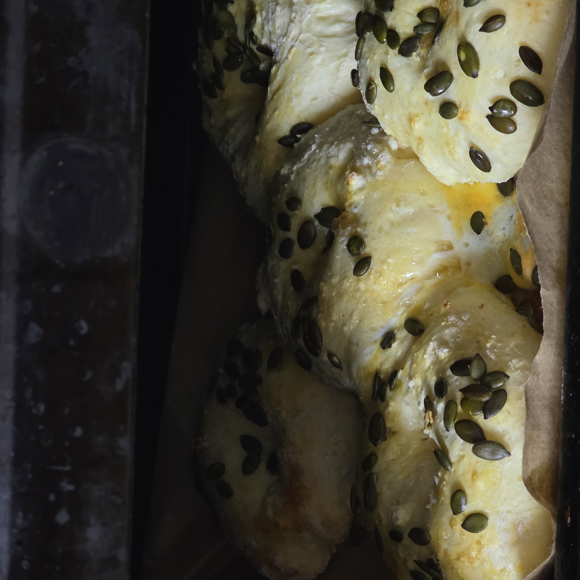 A close-up shot shows a braided loaf of bread, covered in pumpkin seeds, in a baking pan lined with parchment paper. The pan is set inside what appears to be an oven or another container, with a dark interior. The bread has a slightly golden crust. No people are visible. The location is not evident from the image.