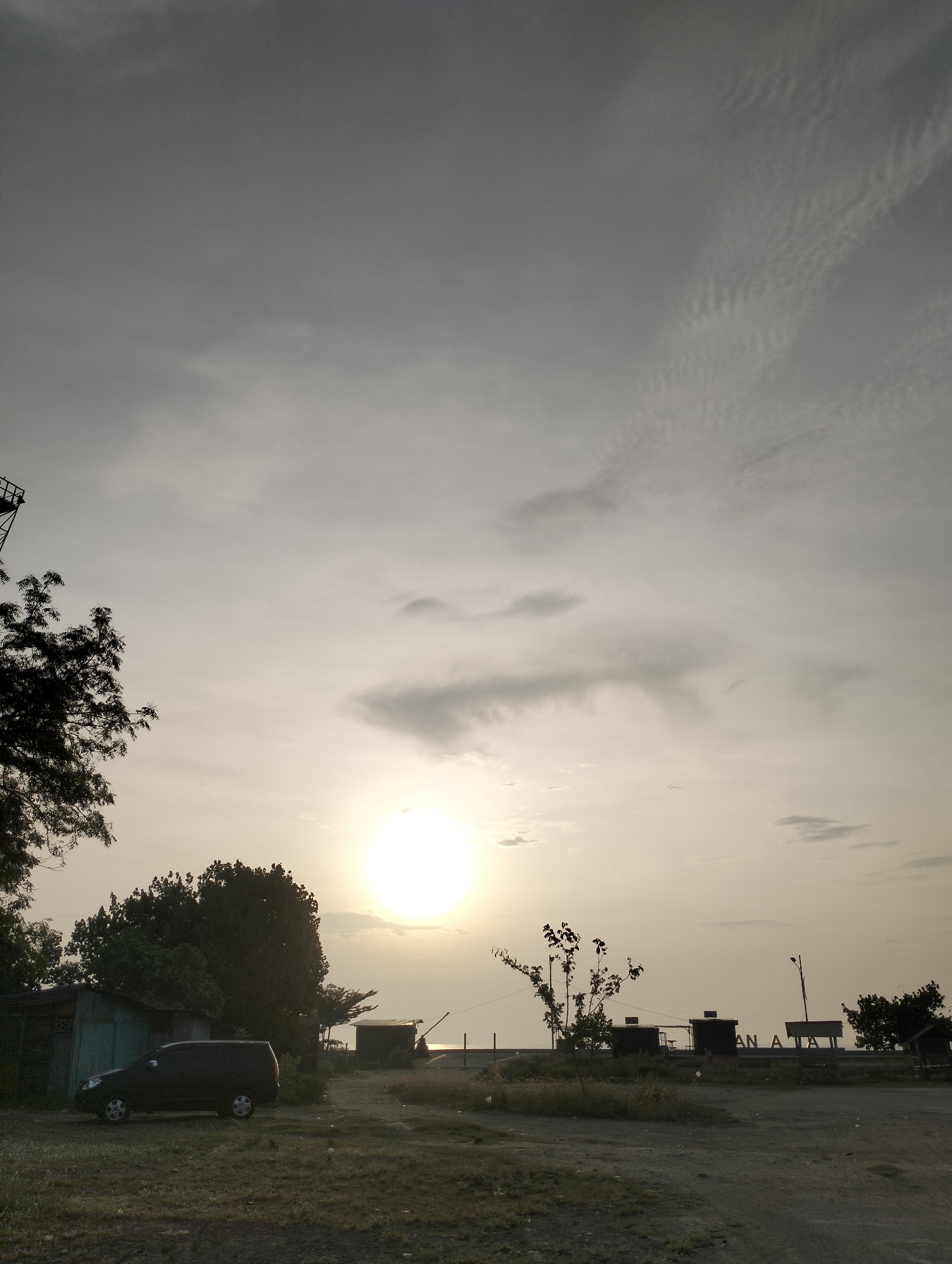 The image portrays an outdoor landscape captured during either sunrise or sunset, indicated by the brilliant, low-hanging sun that dominates the central-right portion of the frame, casting a warm, diffused light across the scene. The sky is largely overcast, featuring a mix of thin, rippled clouds in the upper right and heavier, indistinct clouds elsewhere, creating a soft, hazy ambiance.
The foreground consists of unpaved, dusty ground with sparse patches of grass and low-lying vegetation. On the left, a dark-colored vehicle, possibly an SUV or minivan, is parked beside a small, simple building with a corrugated metal roof. A dense tree with dark foliage stands prominently behind this building. Further into the midground, several more small, rustic structures or kiosks are silhouetted against the bright horizon, situated near what appears to be a body of water, likely the sea, with a visible concrete barrier or embankment. A tall metal structure, resembling a tower, is partially visible on the far left, extending above the treeline. On the right, a streetlight pole stands near the kiosks.
A sign is visible among the structures near the water, displaying the partial text "AN A TA." The scene is devoid of people or overt activity, conveying a sense of quietude and stillness. The overall impression is one of serene contemplation at dusk or dawn in a coastal environment, consistent with a location such as Lhokseumawe, Indonesia.