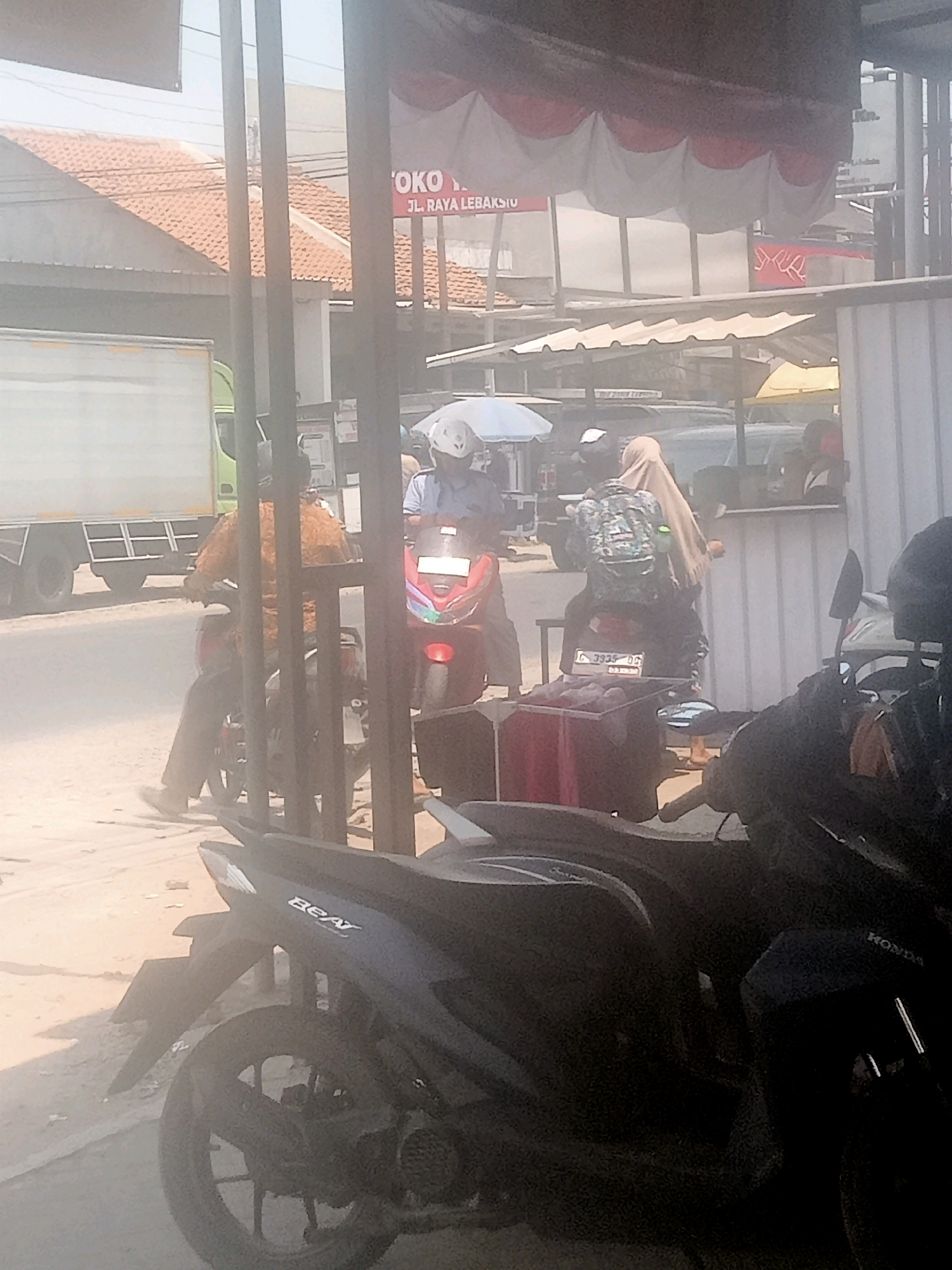 In the city of Lebaksiu, Indonesia, this image captures a street scene with several motorcycles, a truck, and people. In the foreground, a black motorcycle with the "BEAT" logo is parked on a dusty surface. Behind it, another black motorcycle with the "HONDA" logo is partially visible. Further back, two people are riding motorcycles. One person, wearing a helmet and a brown shirt, is on a red motorcycle. Another person, wearing a hijab and a camouflage-patterned backpack, is on a motorcycle with a license plate that reads "C 3935 DG". A large white truck with a green cab is parked on the left side of the image. Overhead, a banner with red and white fabric is draped across a structure, and a sign reads "TOKO JL. RAYA LEBAKSIU". The environment appears to be an outdoor commercial area, possibly a street or market, with buildings and awnings visible in the background. The overall scene is hazy, suggesting a dusty or overcast condition.