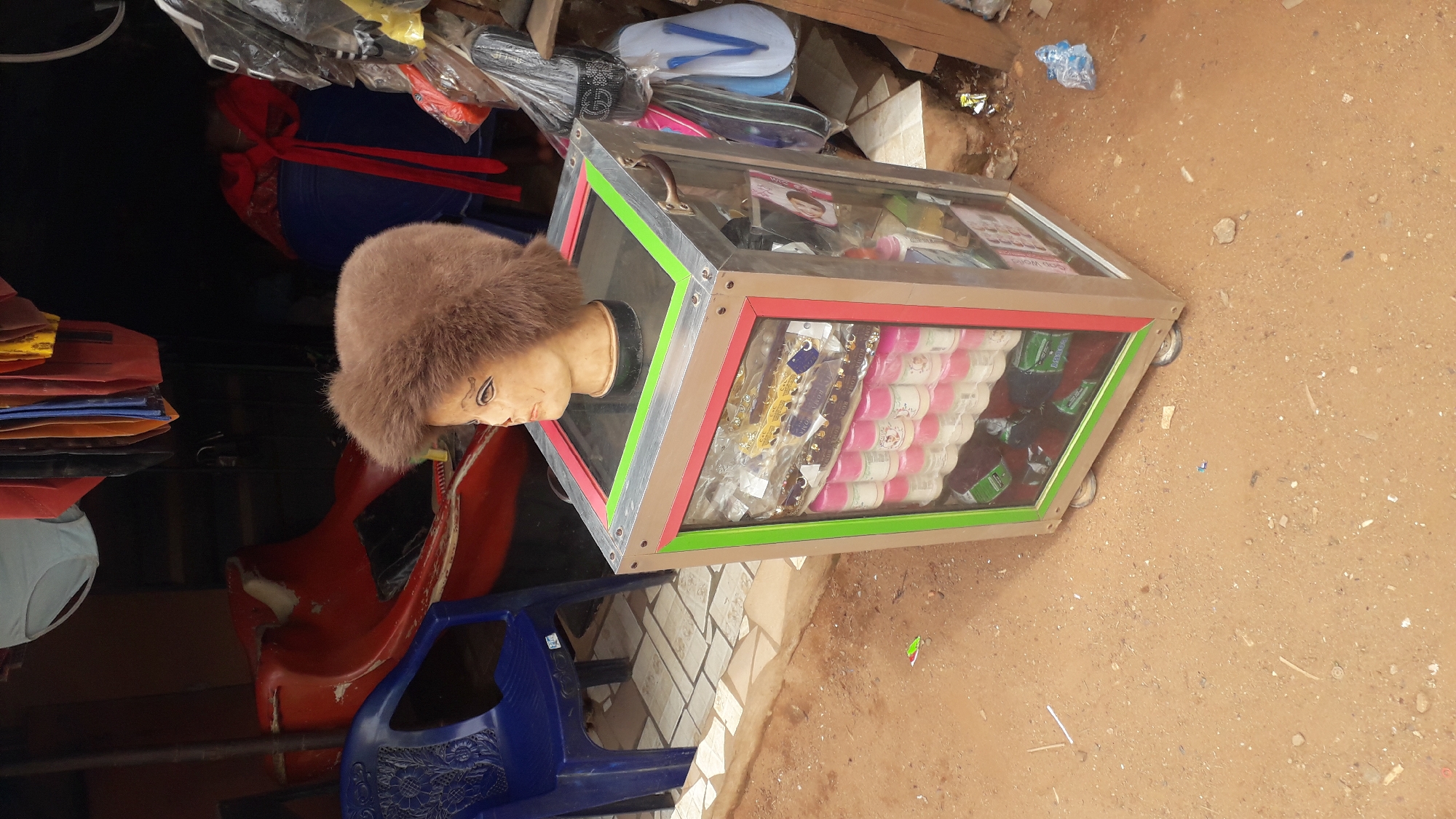 The scene is an outdoor market setting, likely in Onitsha, Nigeria, judging by the style of goods and display. The ground is dirt. There's a makeshift stall or shop visible. A glass display case with a silver-colored frame, accented with red and green trim, rests on wheels. Inside the display case are rows of what appear to be rolls of thread and other small goods. Connected to the display is a mannequin head wearing a fur hat. There is a red plastic slide and a blue plastic chair nearby, surrounded by various merchandise and what seems like trash. There is no one present in the image.