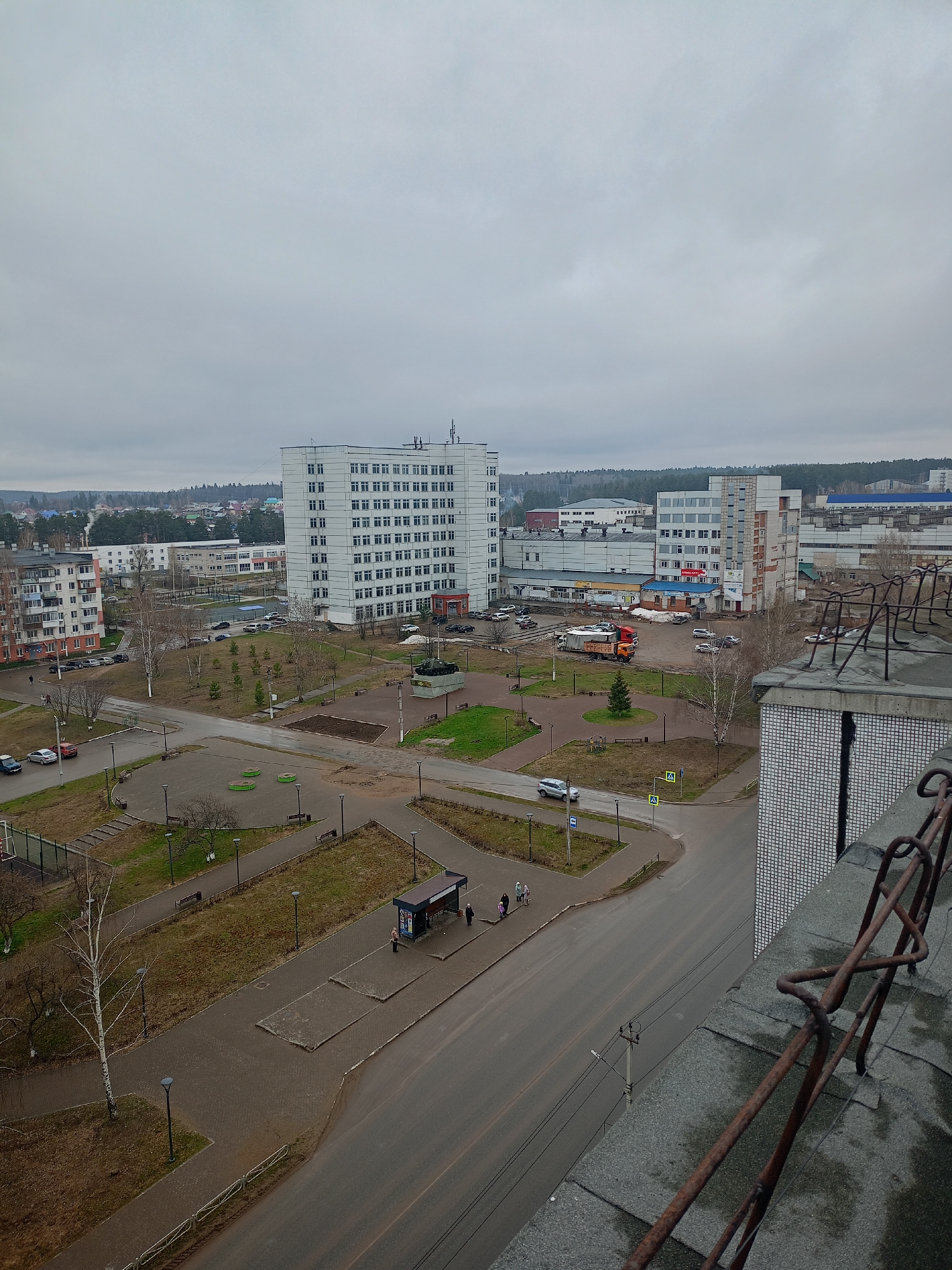 This high-angle photograph presents an urban landscape in Chaikovsky, Russia, under an overcast sky, suggesting late autumn, winter, or early spring due to the bare trees. The scene is viewed from an elevated position, likely a building's upper floor or rooftop, indicated by the rusty railing in the immediate foreground.
At the center, a large, multi-story white building with numerous windows dominates the view, flanked by other apartment blocks and industrial-style structures. To the left, an older residential building features distinctive reddish-orange sections. In the background, a dense forest line marks the horizon.
A central paved square or park area includes walkways, benches, and patches of sparse, brownish grass. A prominent feature is a military tank mounted on a pedestal, serving as a monument. Several vehicles, including parked cars, a white SUV, and a flatbed truck, are scattered across the streets and lots.
A few individuals are visible: people gather at a bus stop on the right and walk along the sidewalks. The roads appear damp, possibly from recent rain. The sky is uniformly grey and cloudy, creating a subdued, diffuse lighting.
Visible text includes what appears to be "АННА РУСЬ" (ANNA RUS) written in Cyrillic on a sign on a building in the mid-right portion of the image. The overall impression is one of a quiet, everyday urban scene.