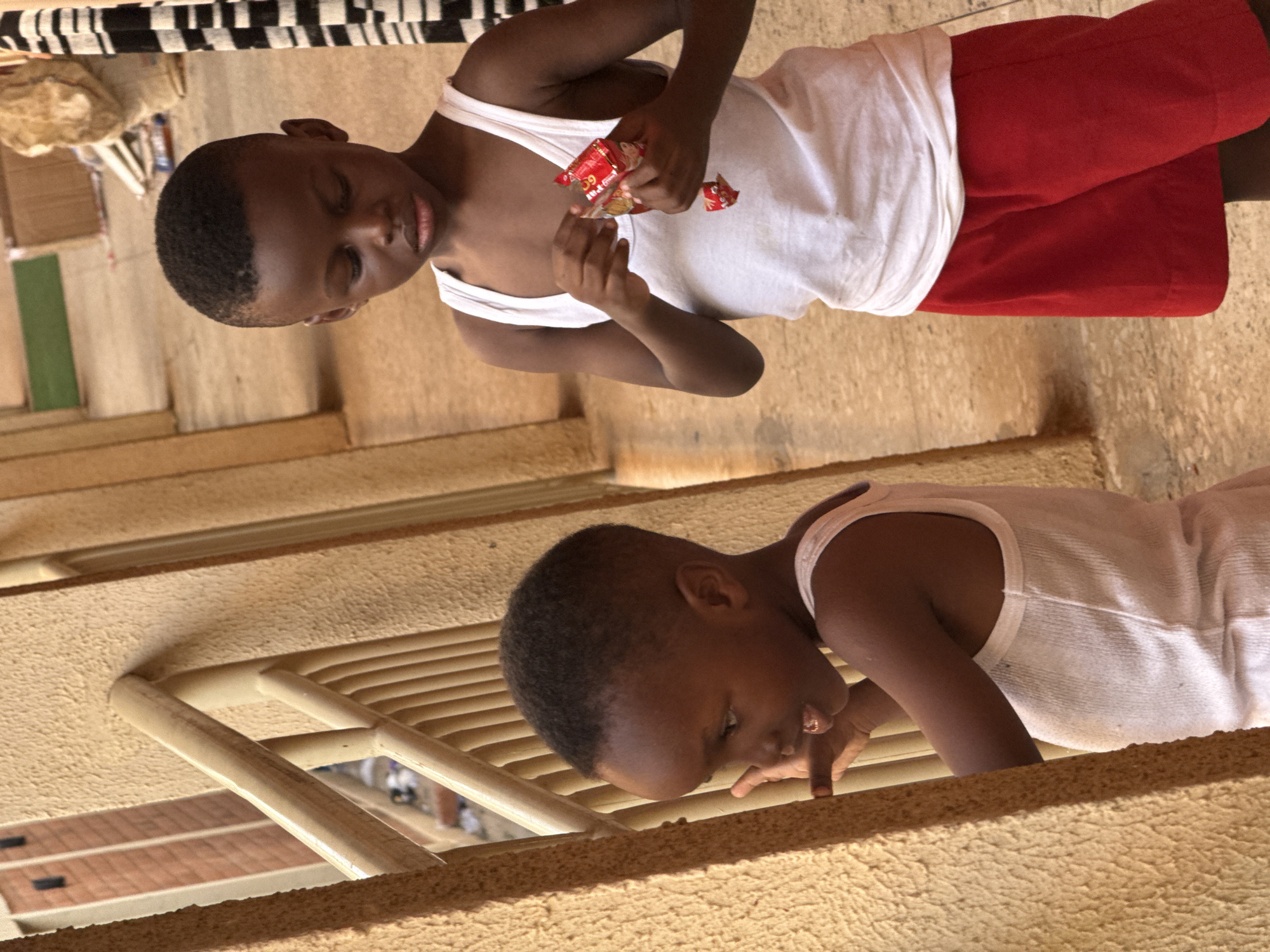 Two young boys are featured in this medium-shot photo. The boy at the top of the frame is wearing a white tank top and red shorts, and is holding pieces of red-and-gold wrapped candy. The boy at the bottom of the frame is wearing a white tank top and beige shorts and is reaching his right hand through what looks like a wooden fence or railing. The location appears to be indoors, with light-colored walls and potentially a tiled floor. The background suggests the city of Abuja, Nigeria.