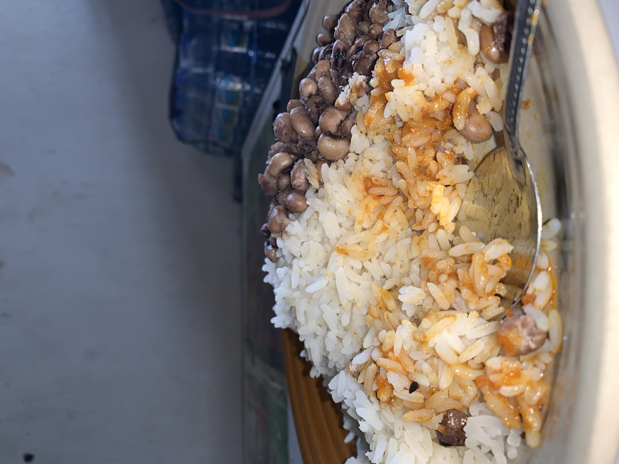 The image shows a close-up of a bowl of food. The food consists of white rice, beans, and a sauce, possibly tomato-based. A spoon is present in the bowl. The location in the city of New Stadium, Nigeria, cannot be confirmed from this image.