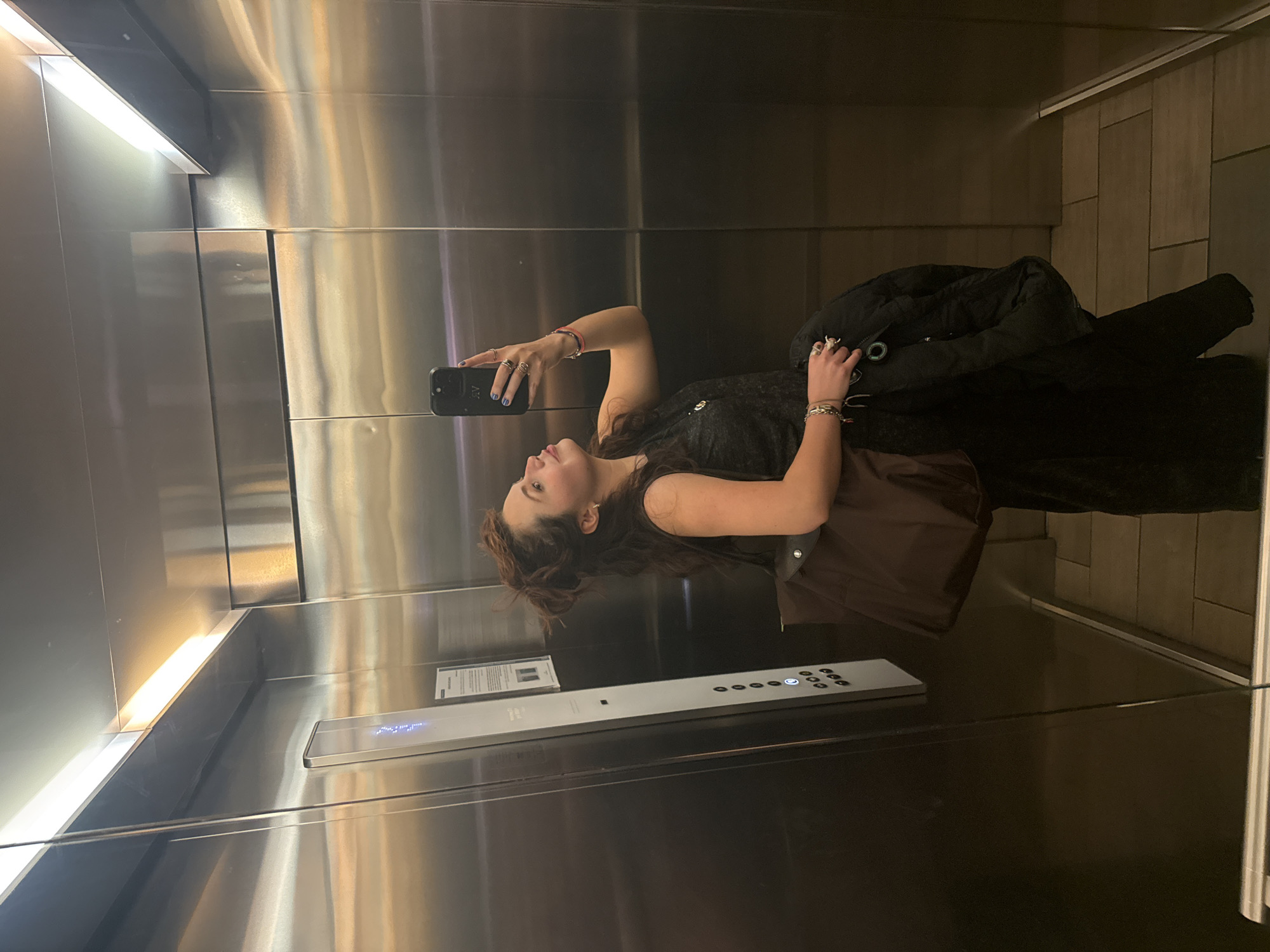 A woman is taking a selfie in an elevator. The elevator has stainless steel walls and a panel of floor buttons. The woman is wearing a dress and has a black coat and brown bag. The location is in Mexico City, Mexico.