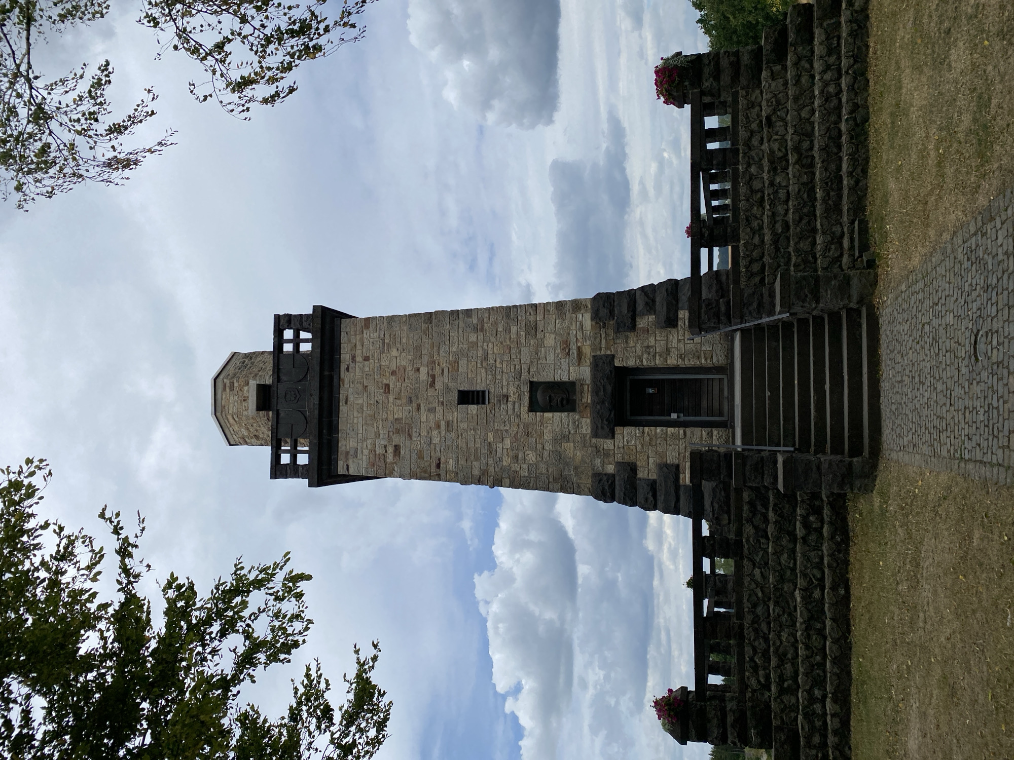 This is a low-angle, full-shot, eye-level, exterior photograph of the Bismarck Tower in Altenkirchen, Germany. The tower is constructed of light-colored stone blocks with darker stone accents. It has a square base that widens at the bottom, a tall, narrow shaft with a small rectangular window, and a top observation deck with decorative stonework and a darker, octagonal cap. Stone steps lead up to a dark wooden door at the base of the tower. A low stone wall with decorative balusters encircles the base of the tower, topped with planters filled with pink flowers on either side of the entrance. Cobblestone pavers lead from the foreground towards the steps. The tower is framed by the branches of trees with green leaves against a cloudy sky. The ground in front of the tower is covered in dry grass with scattered fallen leaves.