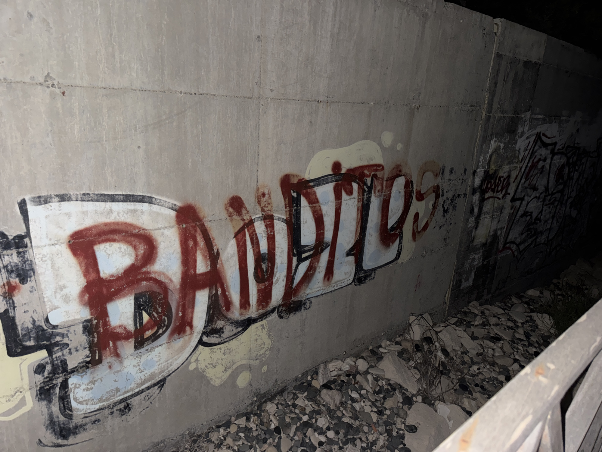 The image depicts a concrete wall covered in graffiti in an outdoor, dimly lit environment, likely an underpass or alcove, near Agios Tychona Community, Cyprus. The graffiti features prominent, stylized lettering in red and black spray paint, with some lighter colored spray paint underneath. The wall itself is a light gray, showing signs of wear and staining. The ground in front of the wall is littered with rocks, gravel, and some dry vegetation. On the right side of the image, a metal railing is partially visible, showing signs of rust. The overall scene suggests an urban or semi-urban, neglected area.