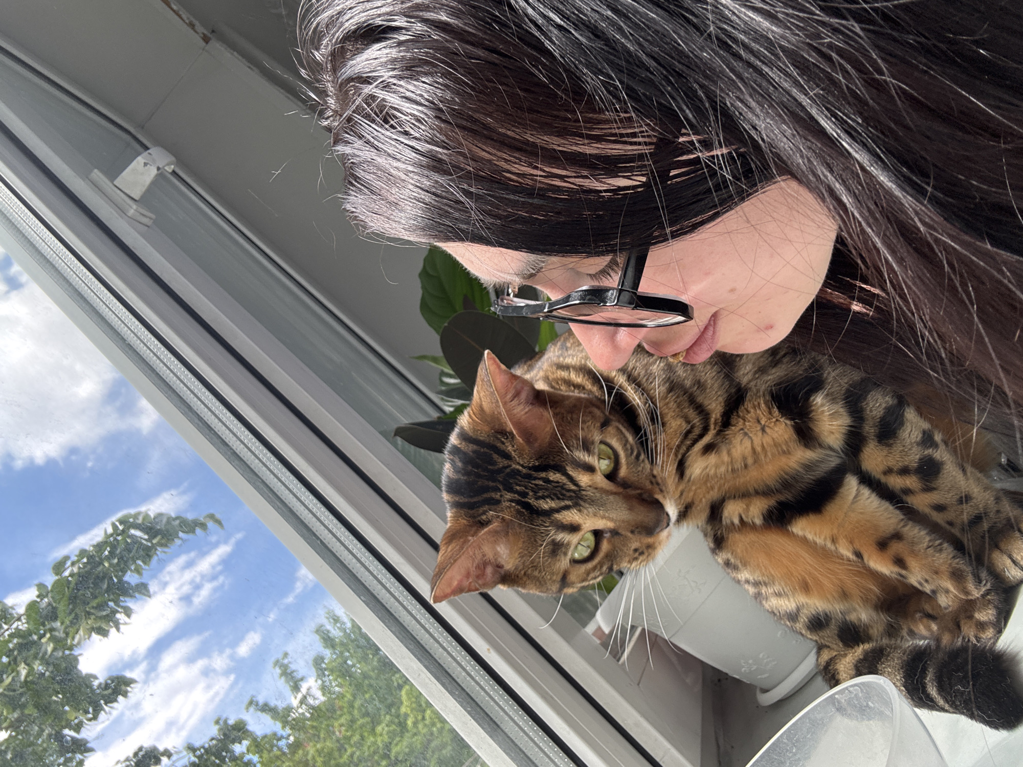 A woman with dark hair and glasses is seen in close proximity to a Bengal cat with a striped and spotted coat. They appear to be near a window with a view of a tree and a sky with patchy clouds. The cat is resting in a white container, possibly a pot, on the window sill.