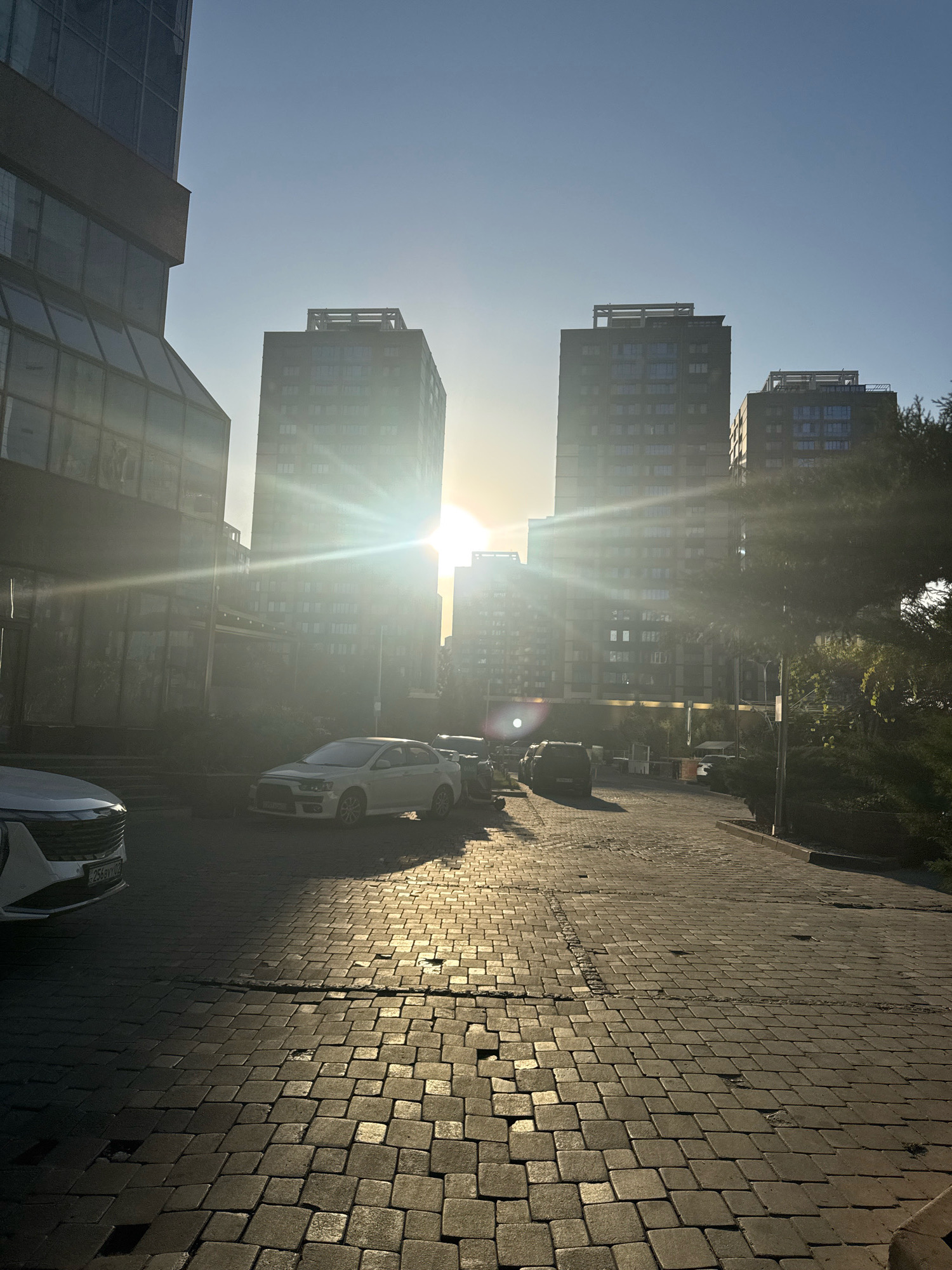 The image depicts a street scene in Almaty, Kazakhstan, during sunset. Two tall, modern buildings dominate the skyline, with the sun's rays creating a strong lens flare effect and casting long shadows. The street is paved with cobblestones, some of which appear uneven. Several cars are parked along the street and on the side. A white sedan is prominent in the foreground, along with the front of another white vehicle. Green trees and bushes line the right side of the street. The sky is clear and blue, with the sun positioned low on the horizon. The overall impression is one of an urban environment bathed in the warm light of the setting sun.