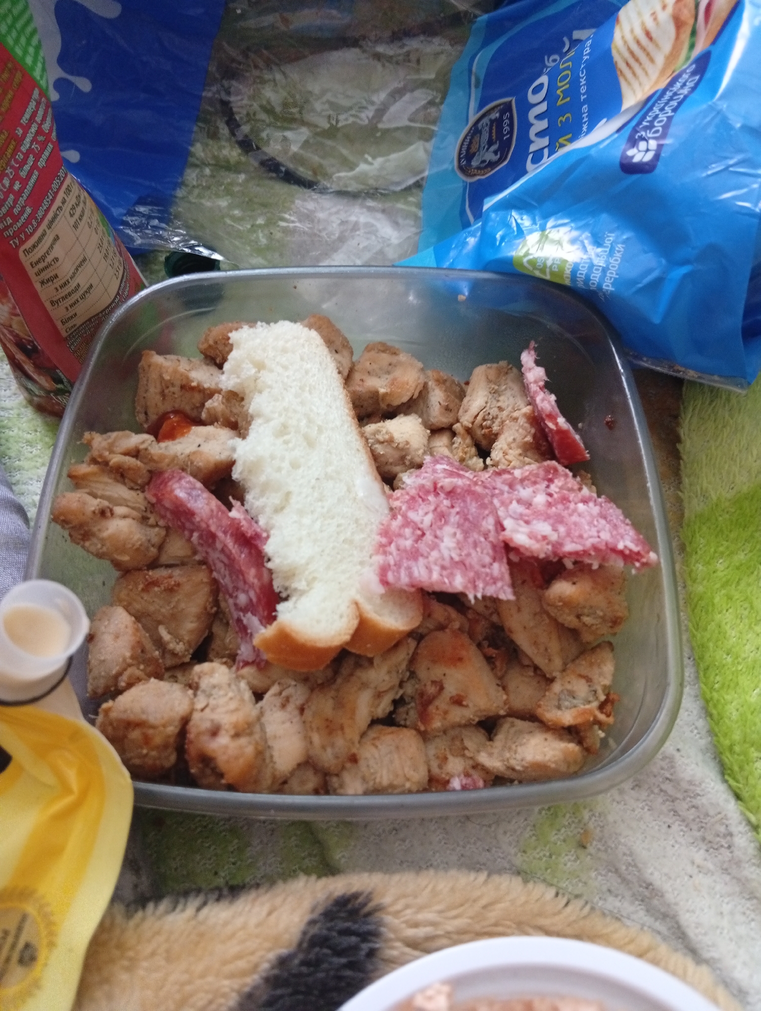 The image shows a plastic container filled with cooked chicken pieces, sliced bread, and sausage. A container of sauce is next to it. In the background are two product packages, one of which is labeled with information in Ukrainian, including the city name "Poltava" and the year "1995." The food appears to be placed on a patterned surface, possibly a towel or blanket.