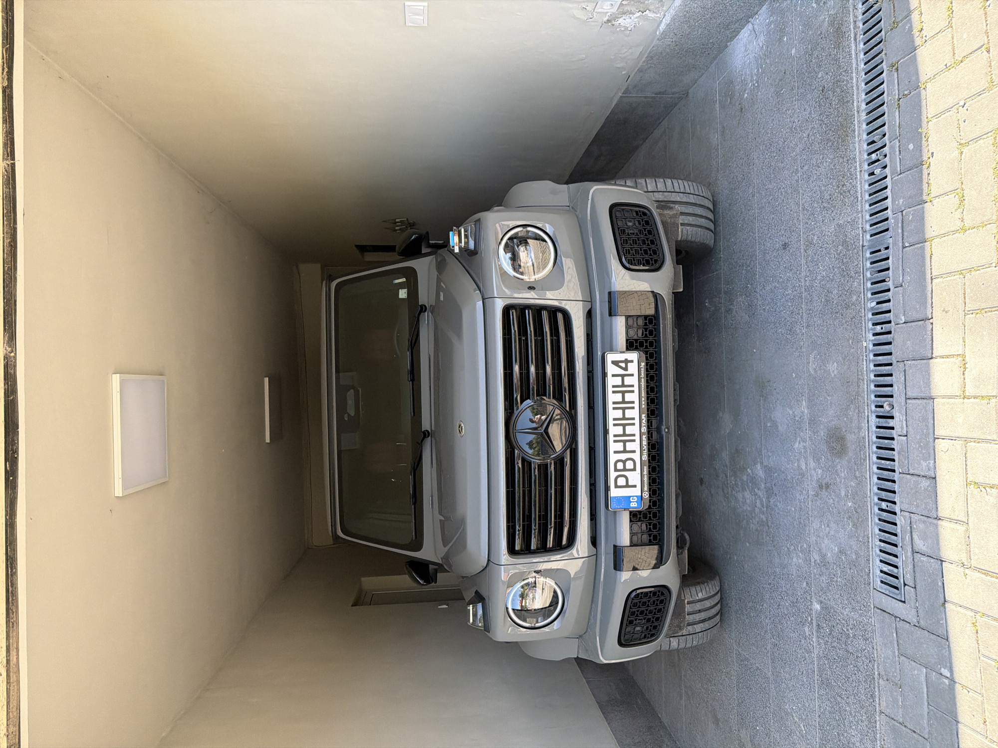 The image shows a gray Mercedes-Benz G-Class SUV parked in what appears to be a narrow alley or garage. The car is positioned facing the camera, with its front grill and headlights prominently visible. The license plate reads "PB HHHHH4" and includes the Bulgarian flag and a Mercedes-Benz logo. The background consists of light-colored walls, suggesting an enclosed space. Outside the garage or alley, a drainage system and pavement are visible. The location is potentially Asenovgrad, Bulgaria, based on the license plate and the website address included on it.
