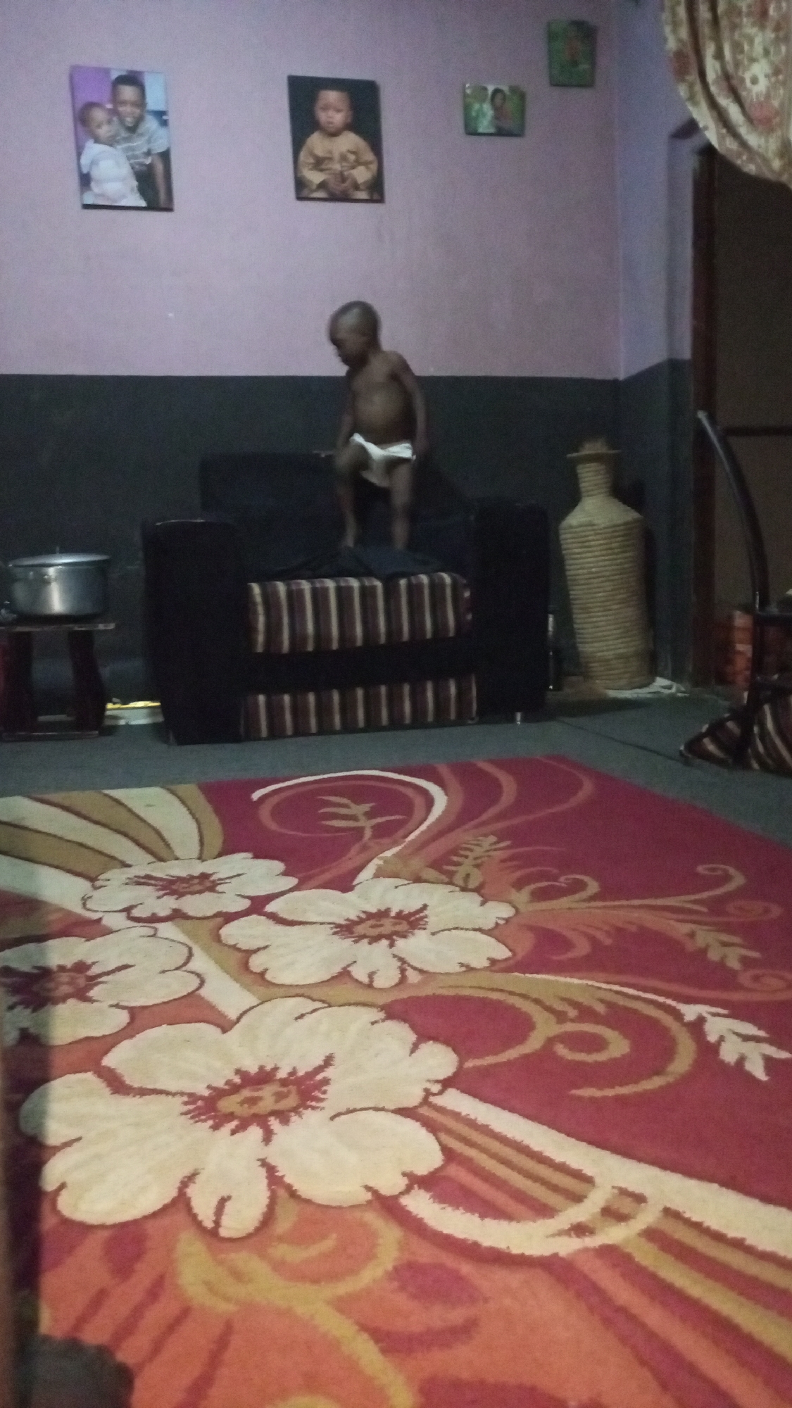 The scene is indoors in Sabon Tasha, Nigeria. A baby wearing a diaper stands on a black and brown striped sofa. Behind the sofa is a wall divided into two colors, the top is lavender and the bottom is black. On the lavender section, three framed pictures hang. To the left of the sofa is a metal pot on a small wooden stool. To the right of the sofa, a tall, woven vase stands beside a dark doorway. The foreground is dominated by a red and cream patterned rug.
