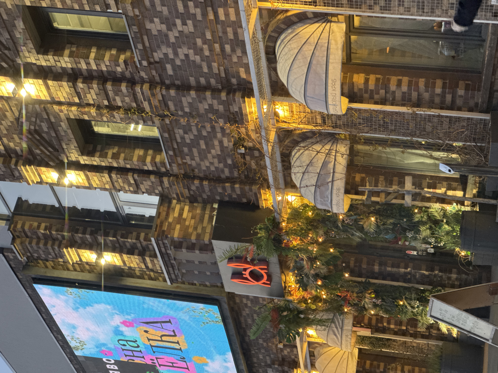 The image shows an exterior view of a brick building in Unknown, Unknown, decorated with Christmas lights and greenery. A large LED screen displays an advertisement in Russian, with the words "на ёлка" (meaning "for the Christmas tree"). Above the screen is an illuminated logo, possibly for a business. The building has windows with awnings, and a metal structure is visible above the awnings. The photograph is taken at night, with artificial lighting illuminating the scene.
