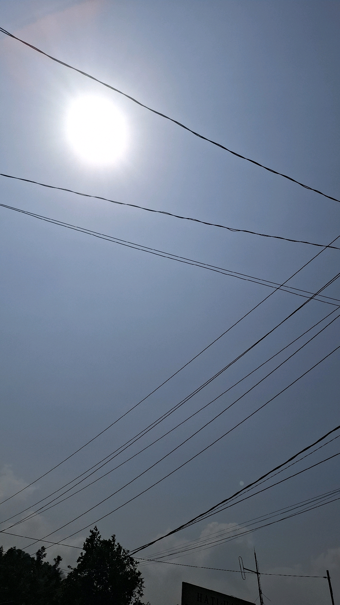 The image shows a bright sun and several power lines against a blue sky. In the lower part of the image, there are silhouetted trees and a sign. The scene appears to be outdoors, possibly in a semi-urban or rural area of Bandar Lampung, Indonesia, given the presence of power lines and trees. The sun is a prominent feature, casting a bright light and creating a slightly hazy effect. The power lines are numerous and run across the frame at various angles, suggesting they are part of the local infrastructure. The trees are dark silhouettes, indicating they are either far away or the light is coming from behind them. The sign in the foreground is partially visible and appears to have some text on it, but it is not legible enough to discern any specific details. The overall impression is one of a sunny day with a clear sky, punctuated by the man-made element of the power lines.