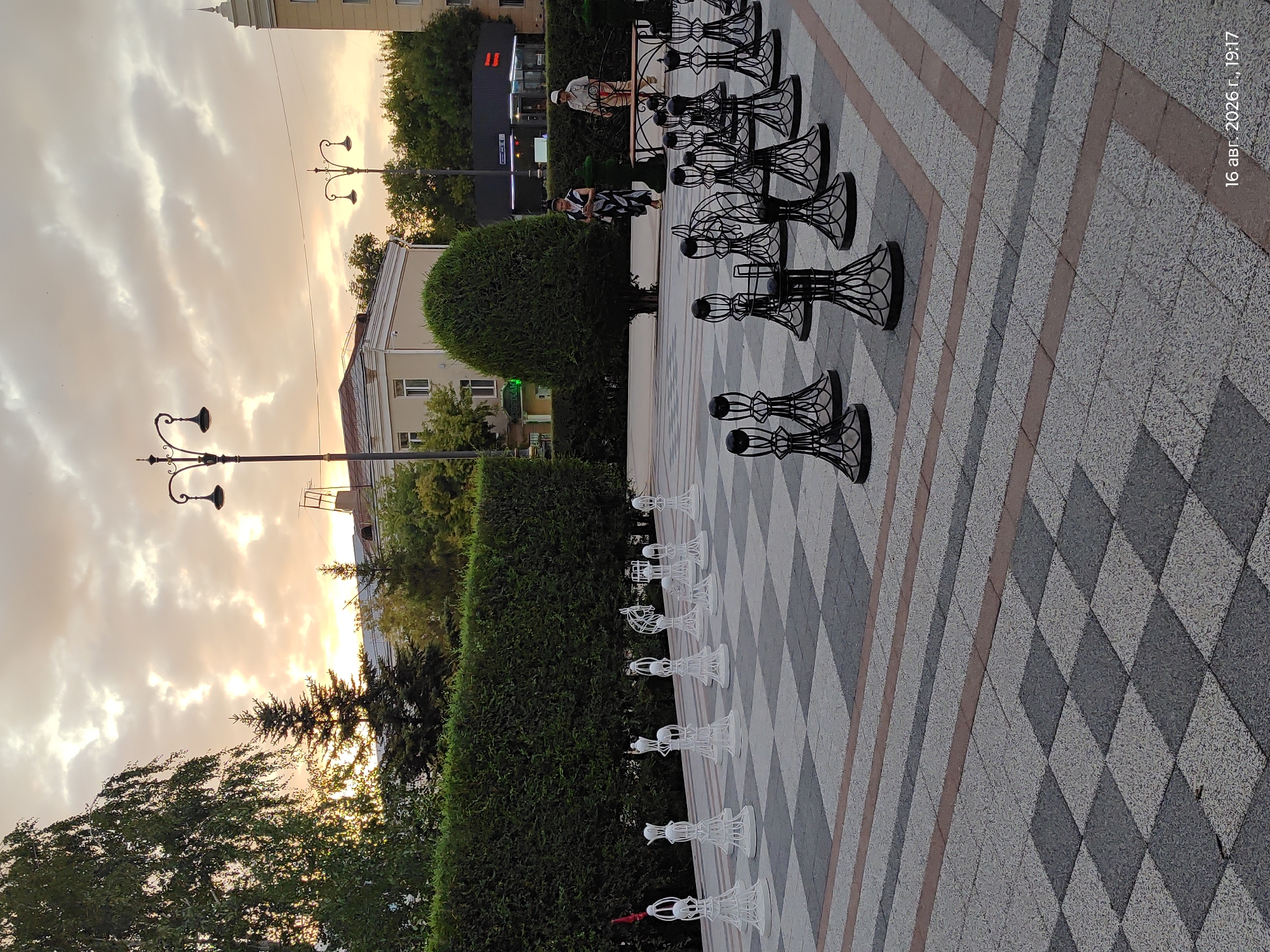 This is a medium shot photograph taken outdoors in what appears to be a public square or park. The foreground features a large outdoor chess set with black pieces arranged on a checkered ground made of grey and white tiles. In the midground, a large, well-maintained green hedge forms a backdrop. Beyond the hedge, two individuals are visible walking on a paved area. The woman on the left is wearing a black and white patterned dress, while the man on the right is wearing a light-colored shirt and trousers, and a hat. A bench is situated behind them, and an intricately shaped green tree stands to their left. Further in the background, buildings and trees are visible under a sky filled with dramatic, cloudy formations during what appears to be either sunrise or sunset, with warm, soft light illuminating the scene. The timestamp in the lower right corner of the image indicates "16 авг. 2026 г., 19:17," which translates to August 16, 2026, at 7:17 PM.