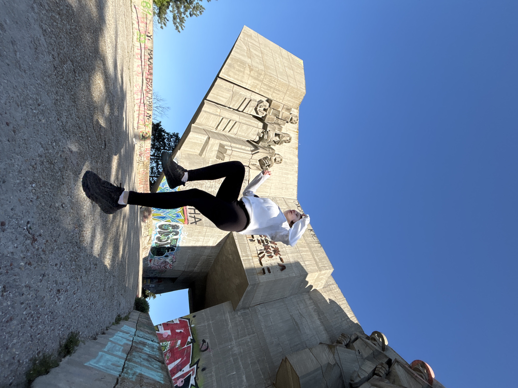 The image displays a creatively oriented outdoor scene featuring a person seemingly levitating against a large brutalist monument. The photograph is rotated 90 degrees counter-clockwise, giving the impression that the ground is a vertical wall on the left and the person is floating horizontally.
The central subject is a young woman, dressed in a white long-sleeved top, black leggings, and dark sneakers. She is captured mid-air, with her body straight and arms bent, hands near her head, creating an illusion of weightlessness or defying gravity. Her expression appears calm and focused.
The setting is dominated by a colossal concrete monument, characteristic of brutalist architecture, which is identified as the Monument to the Bulgarian-Soviet Friendship in Varna, Bulgaria. The monument features several stylized human figures in relief sculptures on its main facade. Below the main structure, an architectural opening or passage is visible. The concrete surfaces of both the monument and the surrounding ground are extensively covered in colorful graffiti and tags, including visible Cyrillic text such as "МАЛКА БЯЛА АРМИЯ" (Small White Army). Patches of trees and sparse foliage are visible in the background and corners of the image.
The scene is illuminated by bright daylight under a clear, vivid blue sky, suggesting sunny weather, likely midday or early afternoon due to the strong shadows cast on the concrete surfaces. The overall atmosphere is artistic and playful, juxtaposing a human figure in an impossible pose against an imposing, graffiti-laden historical structure.