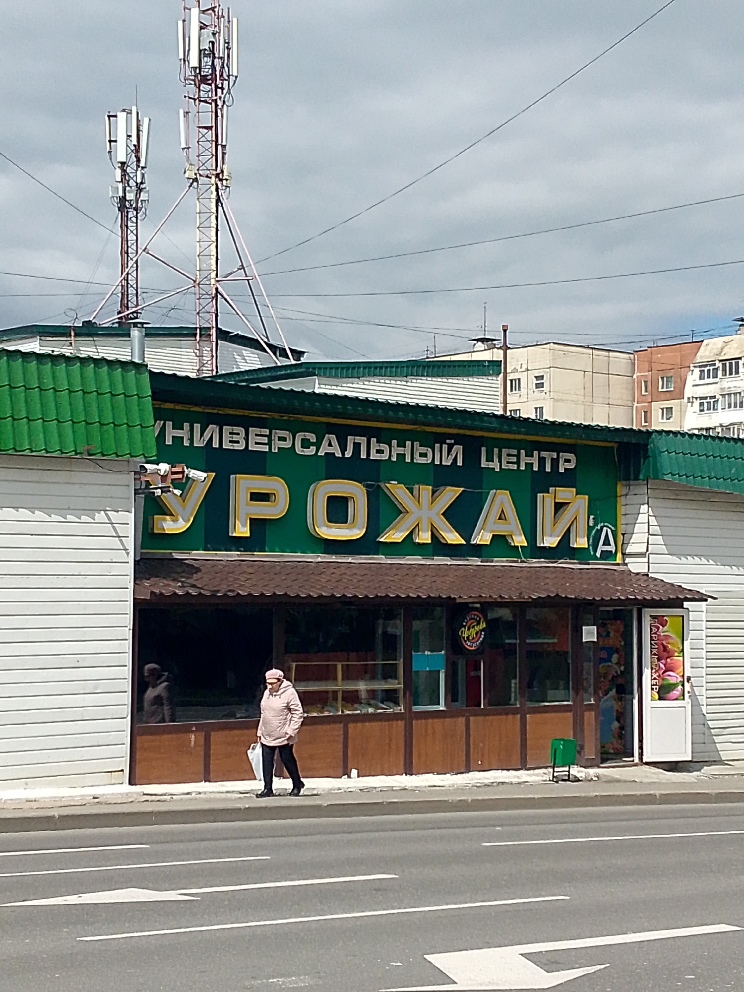 The image shows a street scene in Magadan, Russia, featuring a building with a large green sign that reads "УНИВЕРСАЛЬНЫЙ ЦЕНТР УРОЖАЙ" in large yellow and white letters. Below this sign is a dark brown awning covering the storefront. A woman wearing a light pink jacket and a matching hat is walking on the sidewalk in front of the building, carrying a white plastic bag. The building has white siding and a green corrugated metal roof. In the background, there are tall cell towers and several multi-story buildings. The street in the foreground has white lane markings, including a directional arrow. A green trash can is positioned near the entrance of the building.