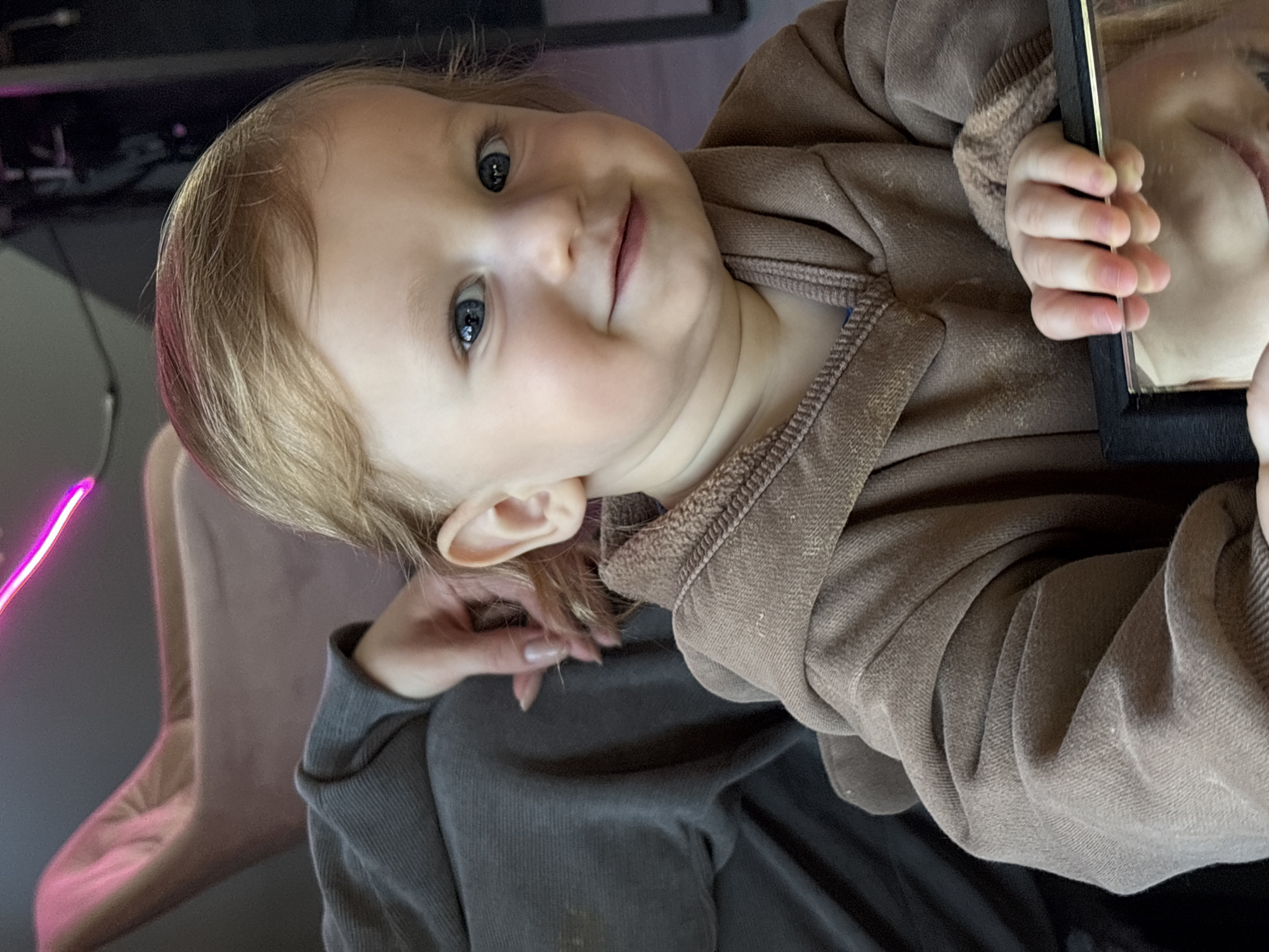 A young child, likely a toddler, is being held or is sitting in someone's lap. The child is wearing a light brown hooded sweatshirt and is holding up a rectangular, framed object, likely a mirror, reflecting a portion of their face. The person holding the child is wearing a dark gray sweatshirt. The background shows a chair, a desk, and a pink neon light. It is not possible to determine the location in Minsk, Belarus, from the image.