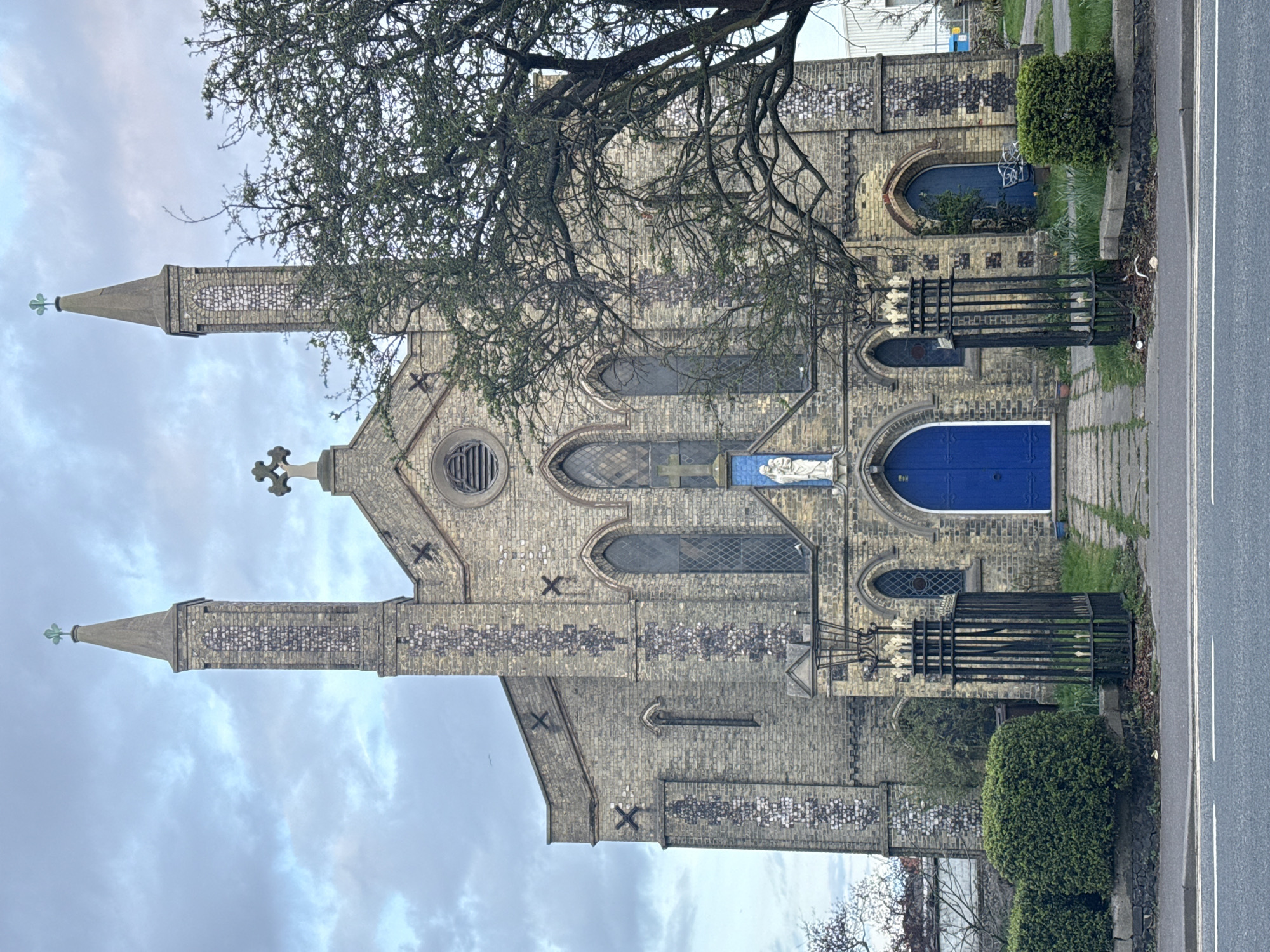 The image shows St. Mary Magdalene Church in Great Yarmouth, United Kingdom. The church is a brick building with two towers and a central cross. The towers are adorned with decorative brickwork. The doors are painted a bright blue. A tree with bare branches partially obscures the view of the upper part of the church. A road runs along the side of the church, with some greenery and a metal structure between the road and the building. The sky is overcast.