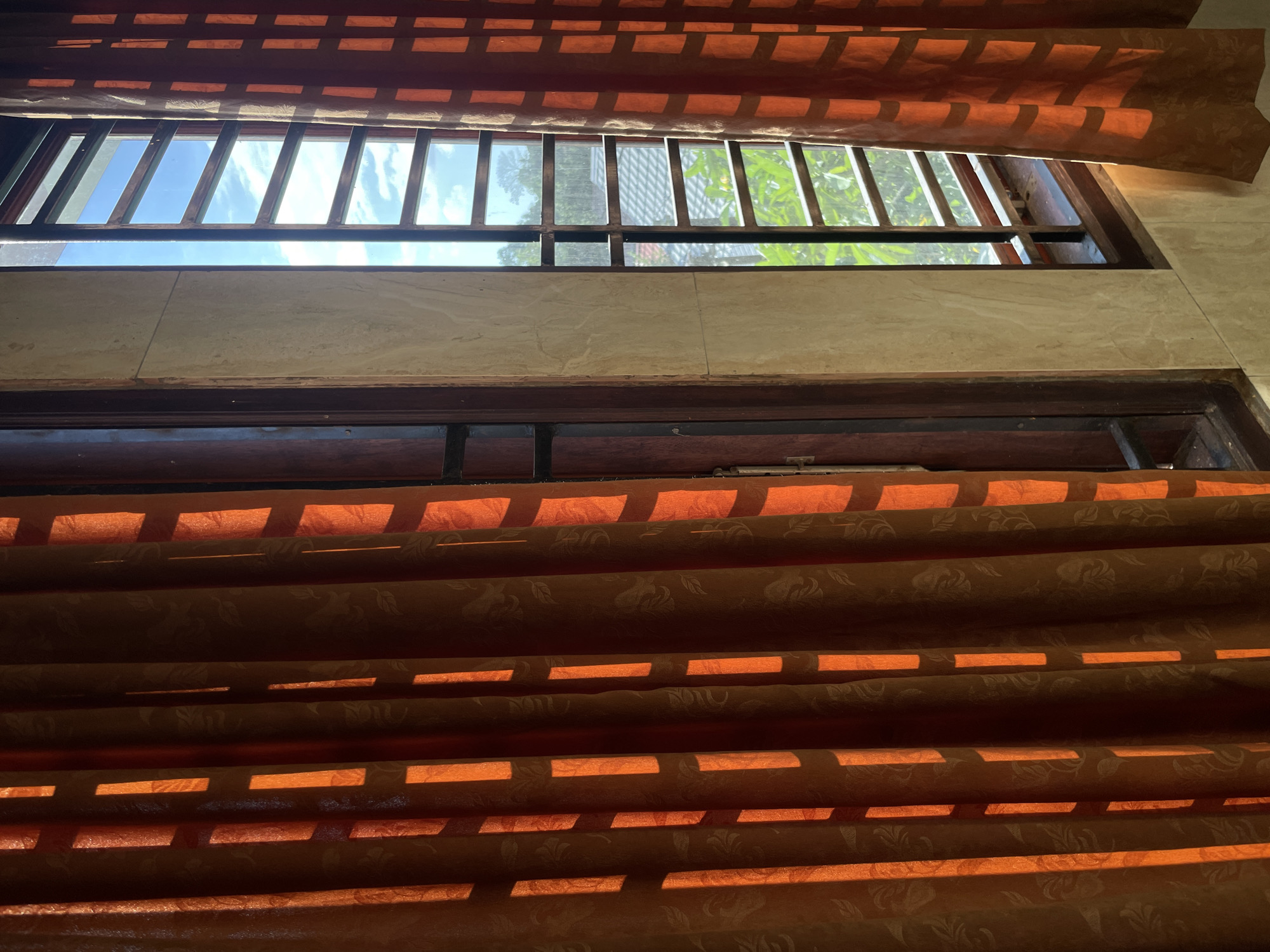 An interior shot captures a window with orange curtains partially drawn, allowing sunlight to cast striped patterns across the fabric. Through the window, a barred view reveals a bright blue sky with scattered white clouds. Outside, glimpses of green foliage and a tiled rooftop are visible. The window frame is dark wood, and the wall beside it is tiled with a light beige, marbelized pattern. The scene appears to be from a residential building in Manado, Indonesia, given the architectural style and outdoor environment, likely in a home or guesthouse.