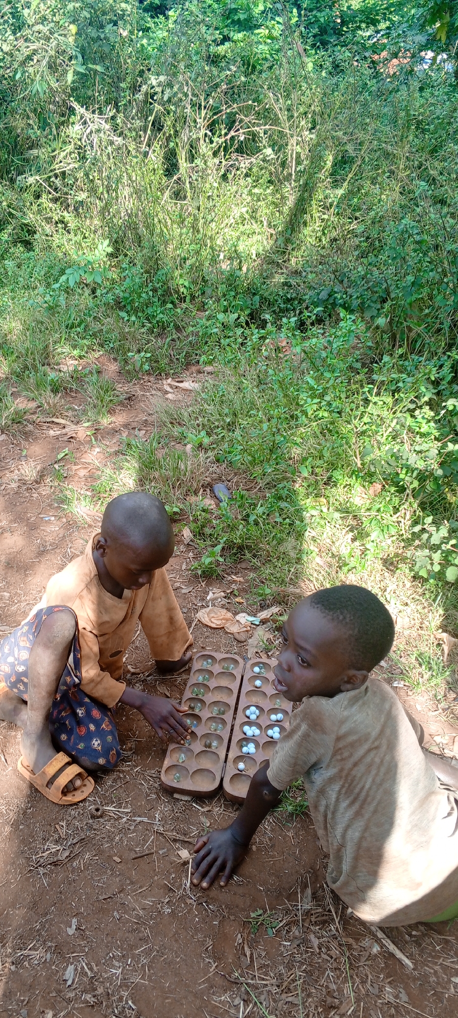Two children play a game with marbles on a wooden game board in Ngalula, Democratic Republic of the Congo. The board sits on the bare ground. One child wears an orange shirt and shorts with soccer ball designs, and sandals. The other child wears a light brown shirt and green shorts. Green vegetation forms the backdrop.