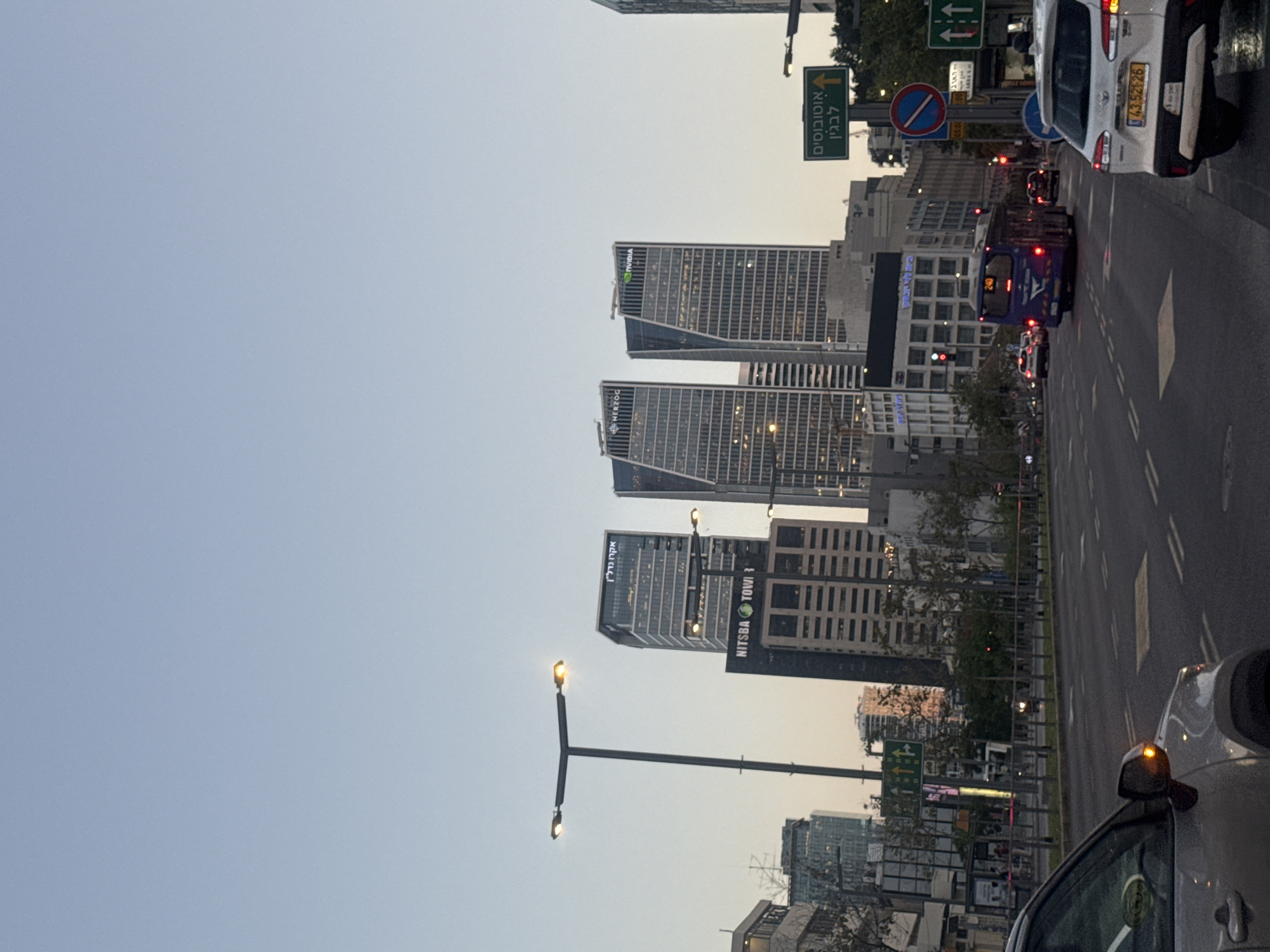 This image depicts an urban street scene in Tel-Aviv, Israel, likely at dusk or early evening, given the light blue-grey sky and the illuminated streetlights and building interiors.
In the mid-ground and background, several modern high-rise buildings are prominent. Corporate logos visible on these structures include "NVIDIA" and "HERZOG". Another tall building displays "NITSBA TOWER" on its side, and another features the Hebrew name "אקרשטיין" (Ackerstein) at its top.
The foreground features a multi-lane road with vehicle traffic. On the far right, part of a white car is visible, showing an illuminated orange turn signal on its side mirror and a license plate reading "43-528-25". Further along the road, a blue public bus, marked with the number "240", has its rear lights on. Other vehicles are also present on the road.
Various traffic signs are visible: a green directional sign points left with Hebrew text "אוטובוסים" (Autobusim - Buses) and "לגבעתיים" (LeGivatayim - To Givatayim). Above it, another green sign indicates straight and right-turn lanes. A "No Stopping" sign and a blue "Mandatory Right Turn" sign are also clearly visible. A street light with two glowing lamps stands in the mid-ground. No people are explicitly visible in the scene.