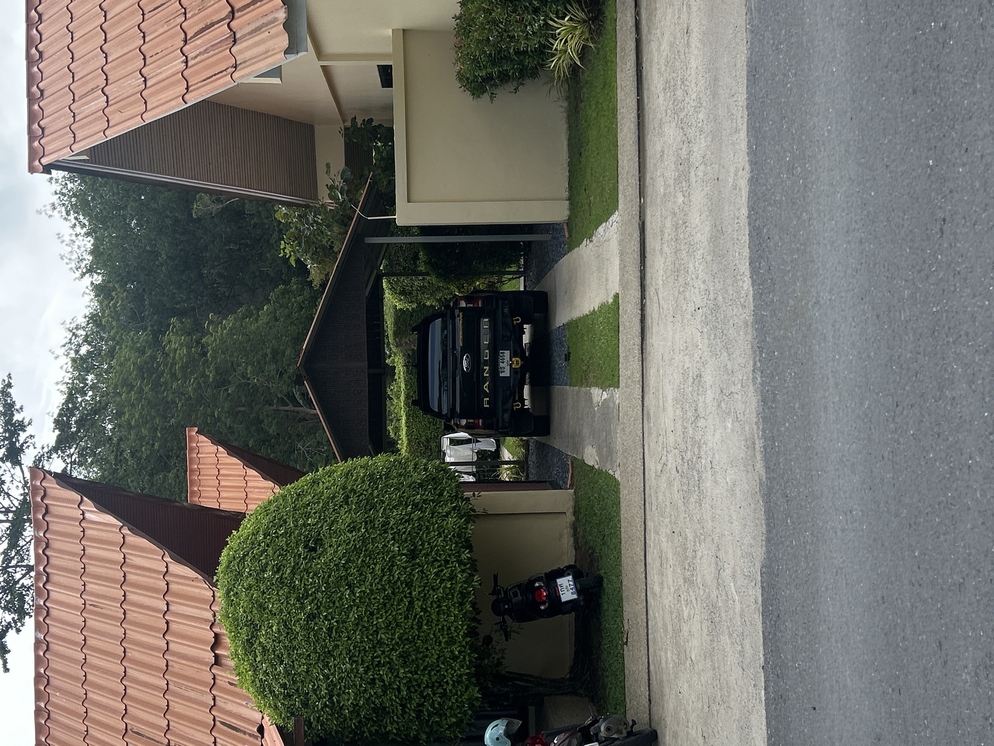 The image shows a street scene in Rawai, Thailand. On the right is a gray asphalt road. To the left of the road is a concrete walkway bordered by grass. A black pickup truck, identified as a "RANGER" by the lettering on the back, is parked in a covered carport. The license plate is partially visible. Behind the truck is a glimpse of another, lighter-colored vehicle. To the lower left of the pickup truck, partially obscured by a large, round, green bush, is a black motorcycle. Further down, more motorbikes are parked and they are partially visible. The houses have terracotta tile roofs. Dense green trees are visible in the background. The weather appears overcast.