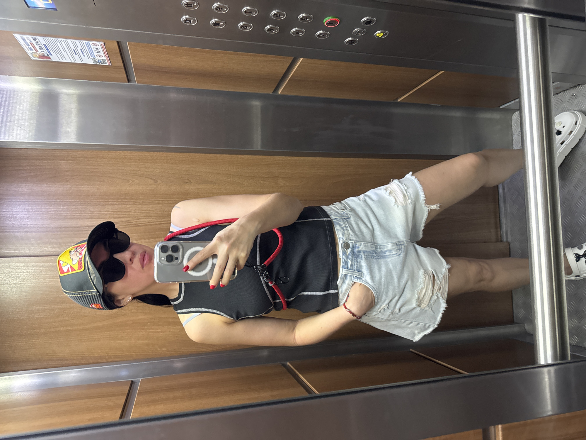 A woman stands in an elevator, taking a selfie in the mirror. She is wearing a black tank top with white stitching, light blue distressed denim shorts, a black baseball cap with a yellow and red graphic, and sunglasses. A red cord is attached to her top and draped over her shoulder. She has red nail polish on her fingernails. In her right hand, she holds a smartphone. Her left hand is in her pocket. The elevator has wood-paneled walls and a brushed metal control panel with buttons. The floor of the elevator is metallic. A poster with Russian text is visible on the wall behind her. The image appears to be taken in a building in Barnaul, Russia.