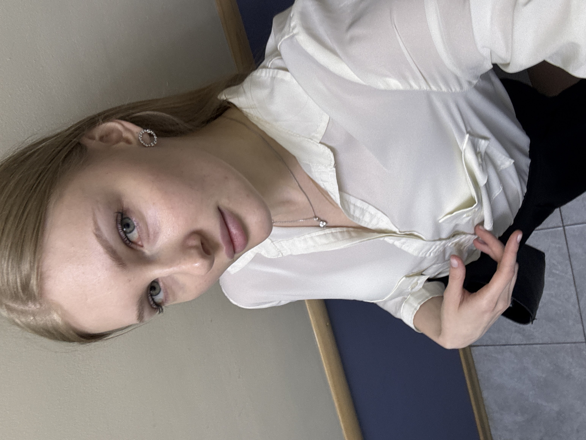 A woman with light blonde hair and blue eyes, wearing a white blouse and a silver necklace with a heart-shaped pendant, poses indoors. She has a round earring in her left ear. The background shows a wall with a beige section and a blue section separated by a wooden trim. The floor appears to be tiled. The subject is holding a black garment or item with her right hand. The location is implied to be in Perm, Russia, but there is no visible evidence of this in the image.