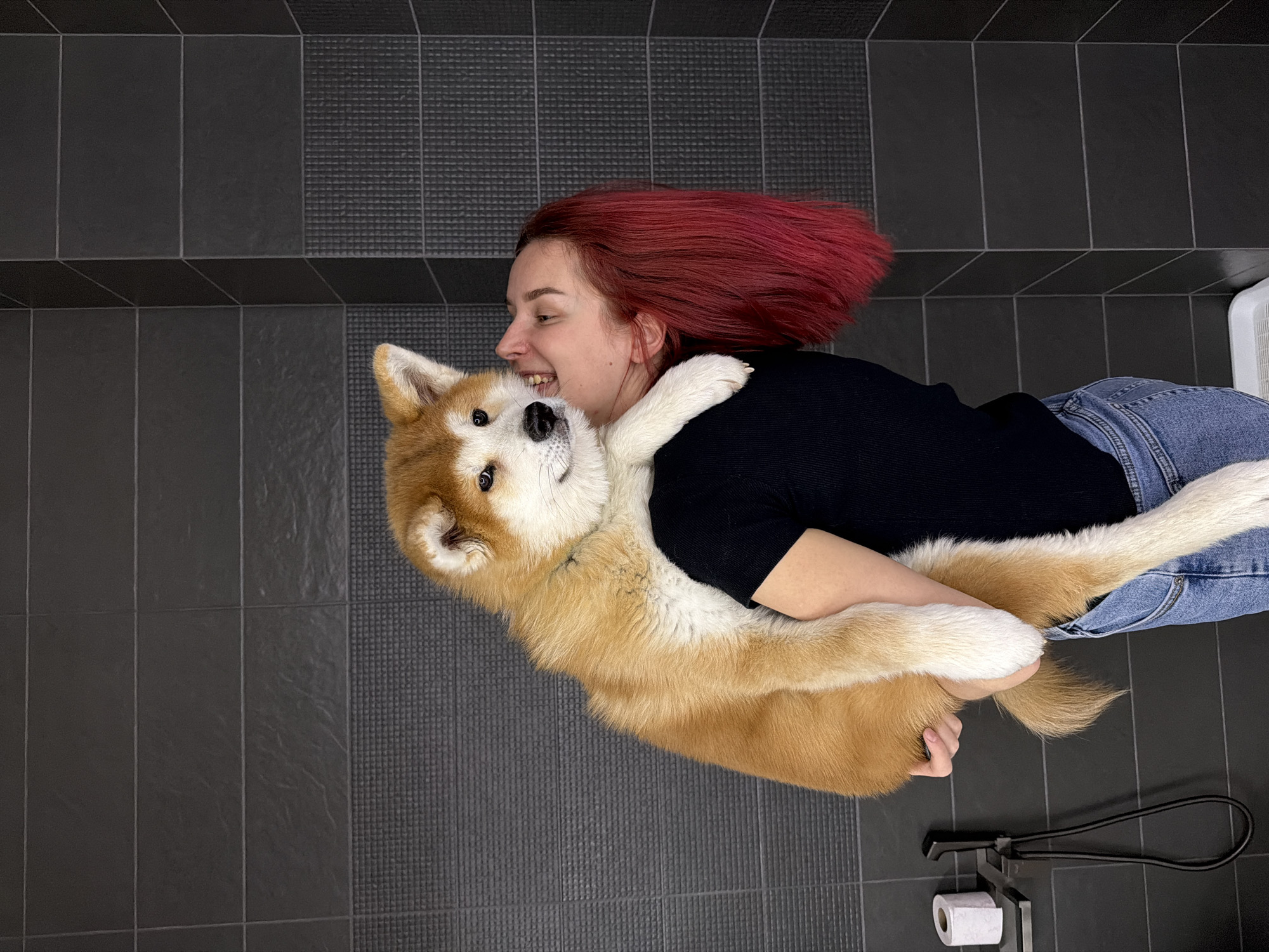 An overhead shot captures a joyful moment between a woman and a dog lying on a dark grey tiled floor, possibly in Chaikovsky, Russia. The woman, with vibrant red hair, is positioned on her side, wearing a black long-sleeved top and blue jeans. She is smiling broadly, with her head resting close to the dog's head, and her arms are wrapped around the dog in an affectionate embrace.
The dog, an Akita with light brown and white fur, is also on its side, facing the camera with a happy, somewhat playful expression. Its front paws are around the woman's neck/shoulder, mirroring the hug. Both subjects appear deeply content and connected.
The floor is made of large, rectangular dark grey tiles. Some sections of the floor feature a textured, possibly non-slip, pattern, while others are smooth. In the bottom right corner, a black metal toilet paper holder with a white roll of toilet paper is visible, suggesting the setting might be a bathroom or a utility room. The lighting appears even, suggesting an indoor setting without direct sunlight. There is no visible text in the image.
