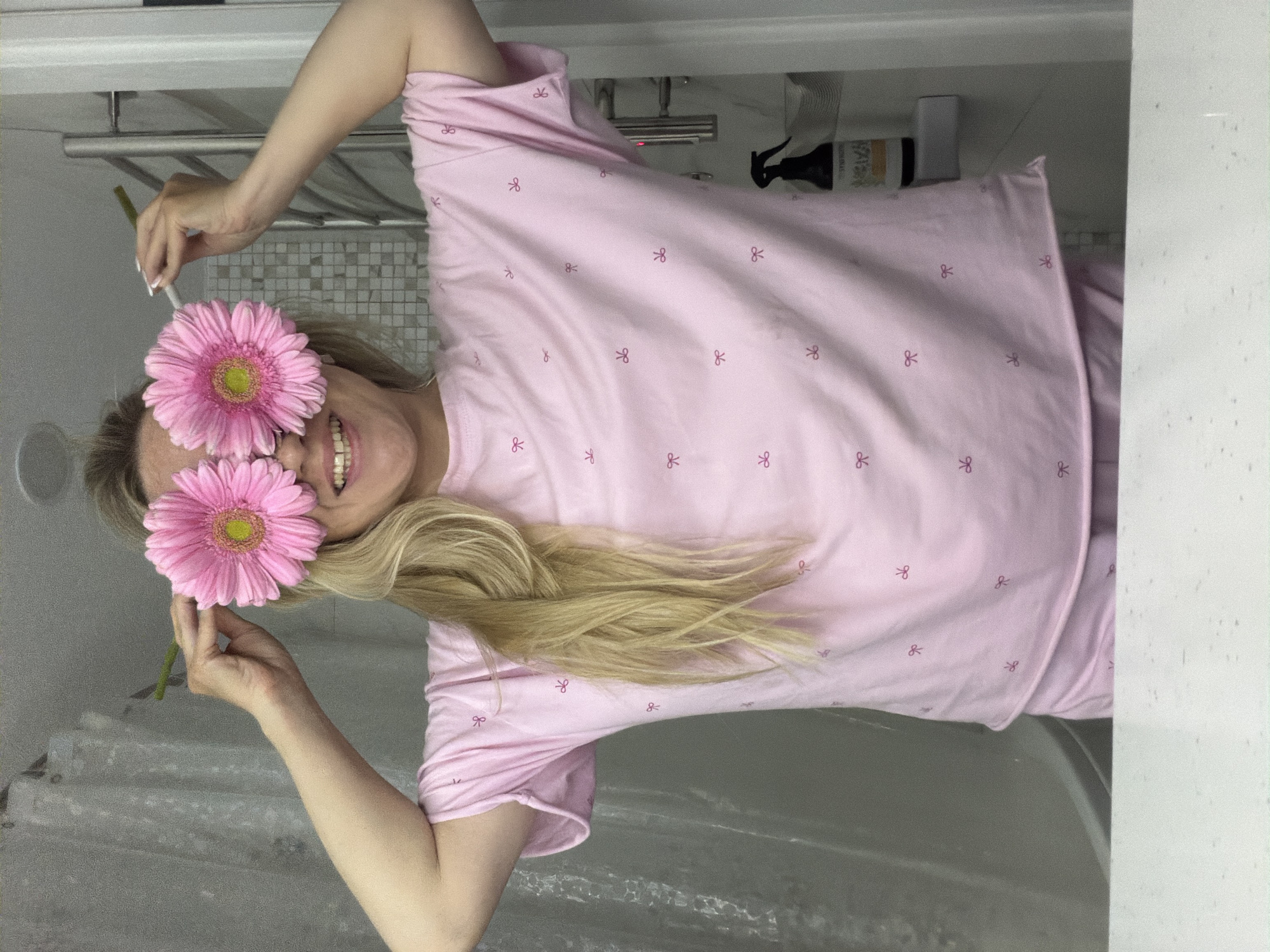 The image shows a woman in a pink pajama top with a bow pattern, smiling with two pink gerbera daisies held over her eyes. She has long, blonde hair draped over the fabric of her pajama top. The background suggests a bathroom setting, with a towel rack, shower curtain, and various bottles visible. There is no indication that this image was taken in the city of Votkinsk, Russia.