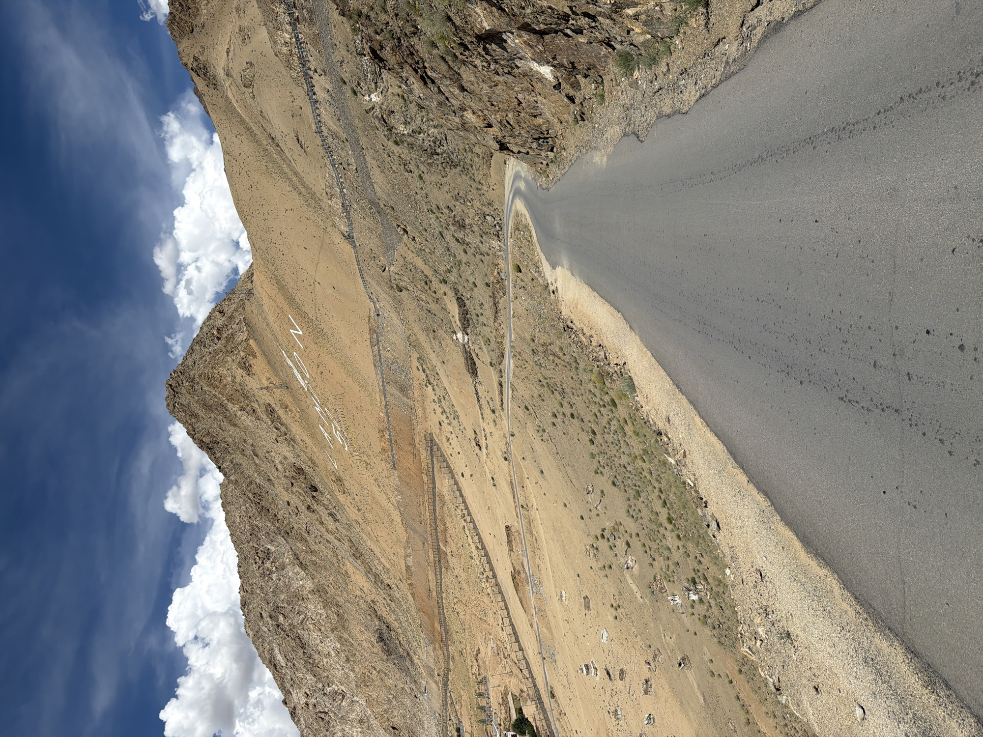 The image depicts a winding asphalt road ascending a steep, arid mountain slope in Leh, India. The terrain is characterized by dry, sandy soil and sparse vegetation. On the mountainside, white letters spell out "LEH" in large block capitals. A wooden fence runs along the side of the road in several sections, presumably to prevent rockfalls or to delineate the path. In the distance, a few small, scattered structures can be seen on the lower slopes, appearing to be stupas or chortens, common religious monuments in the region. The sky is a vibrant blue, dotted with fluffy white clouds. The overall impression is one of a remote, high-altitude landscape, typical of the Himalayan region.