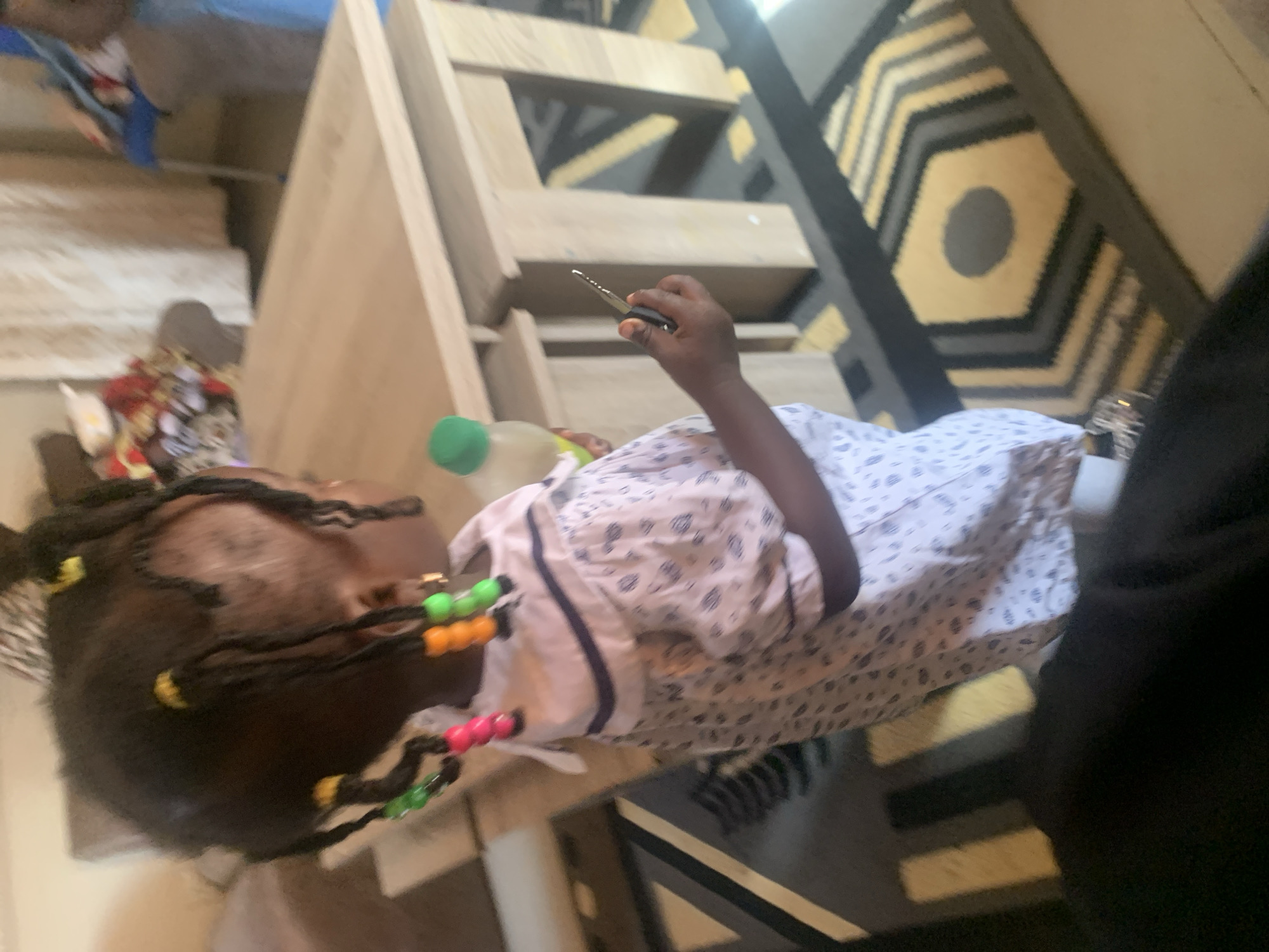 In Gura Top, Nigeria, a young girl with braided hair adorned with colorful beads holds a plastic bottle and a car key in her right hand. She is wearing a light-colored, patterned dress with a dark blue collar. The girl stands in front of a light-colored wooden table, part of which is visible. The flooring is a rug with a geometric pattern in shades of yellow, grey, and black. In the background, curtains are drawn, and a blue chair is partially visible. The overall scene suggests an indoor domestic setting.