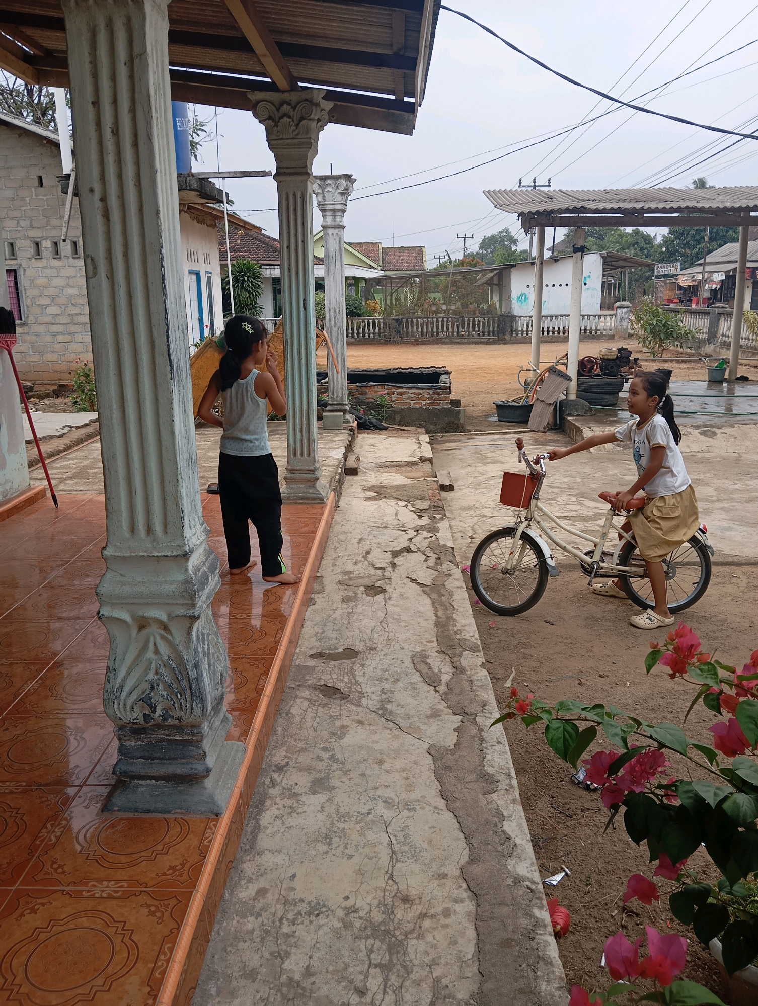 In the city of Leman Benawi, Indonesia, a girl with dark hair tied in a ponytail, wearing a light-colored tank top and dark pants, stands on a tiled patio in front of a white pillar. She faces away from the camera, looking towards a house. Nearby, another girl, also with dark hair in a ponytail, sits on a white bicycle. She wears a white t-shirt and a tan skirt, and her left hand rests on the handlebars. The bicycle has a red basket attached to the front. The background shows a rural or semi-rural environment with various structures, including houses, a covered area with a corrugated roof, and some trees. The ground is a mix of dirt and concrete pathways. Pink bougainvillea flowers are visible in the foreground and to the right of the frame. The lighting suggests it is daytime.