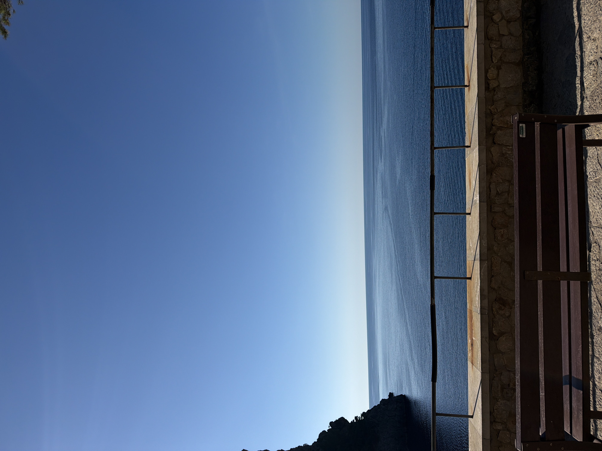 The image shows a view from a stone balcony overlooking the Mediterranean Sea in Escorca, Spain. The sky is clear blue on the left, transitioning to the dark blue of the sea on the right. A metal railing runs along the edge of the balcony, with a wooden bench on the right. Some rocky, vegetation-covered land is visible in the lower center of the image.