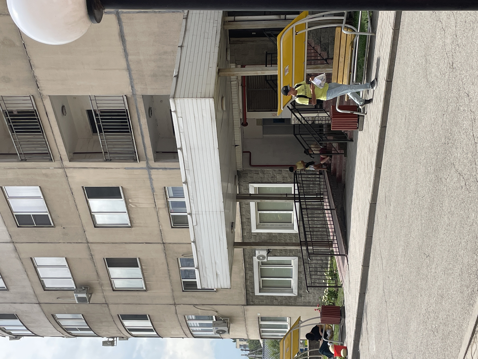 The image shows a residential building in Almaty, Kazakhstan. A young man in a yellow t-shirt and blue jeans walks along a sidewalk in front of the building, holding a white bag and looking at his phone. To his left, a person sits on a yellow bench under a yellow awning, and to the far left, a red bucket and some large round objects are visible. A ramp with a metal railing leads up to the building entrance, where two children are visible on the steps. The building is a multi-story structure with cream-colored facade and numerous windows. A large, white globe-shaped lamp stands on the right side of the frame. The overall scene depicts an urban streetscape on a bright day.